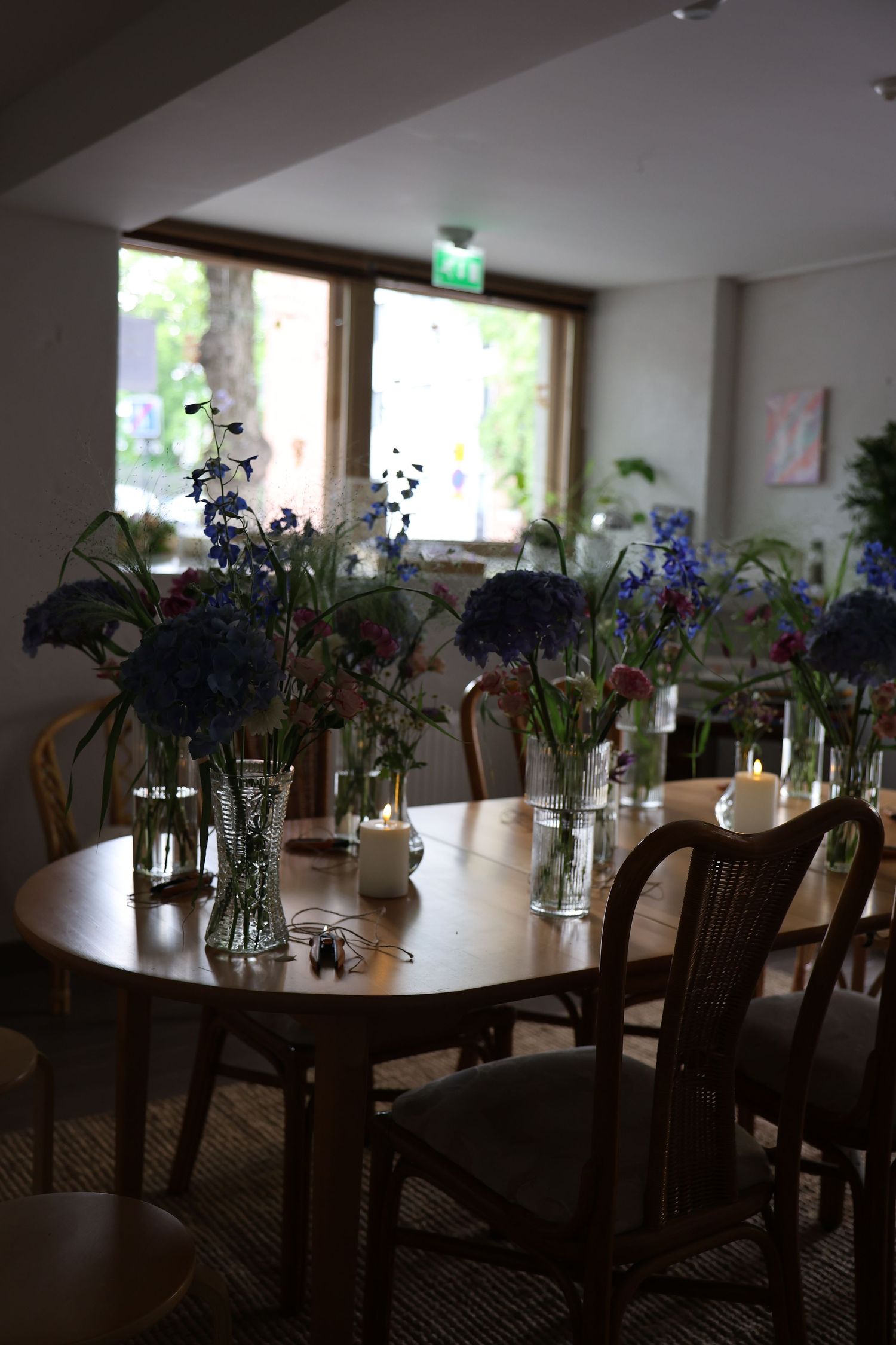 Cozy cafe interior with wooden tables and chairs adorned with blue hydrangea and wildflower arrangements near a bright window.
