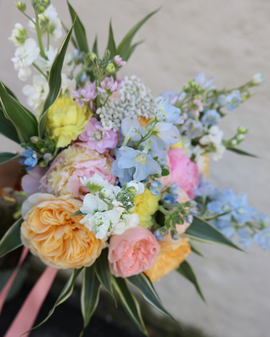 Colorful spring wedding bouquet with peach garden roses, pink peonies, blue delphinium, yellow ranunculus, and white blooms.