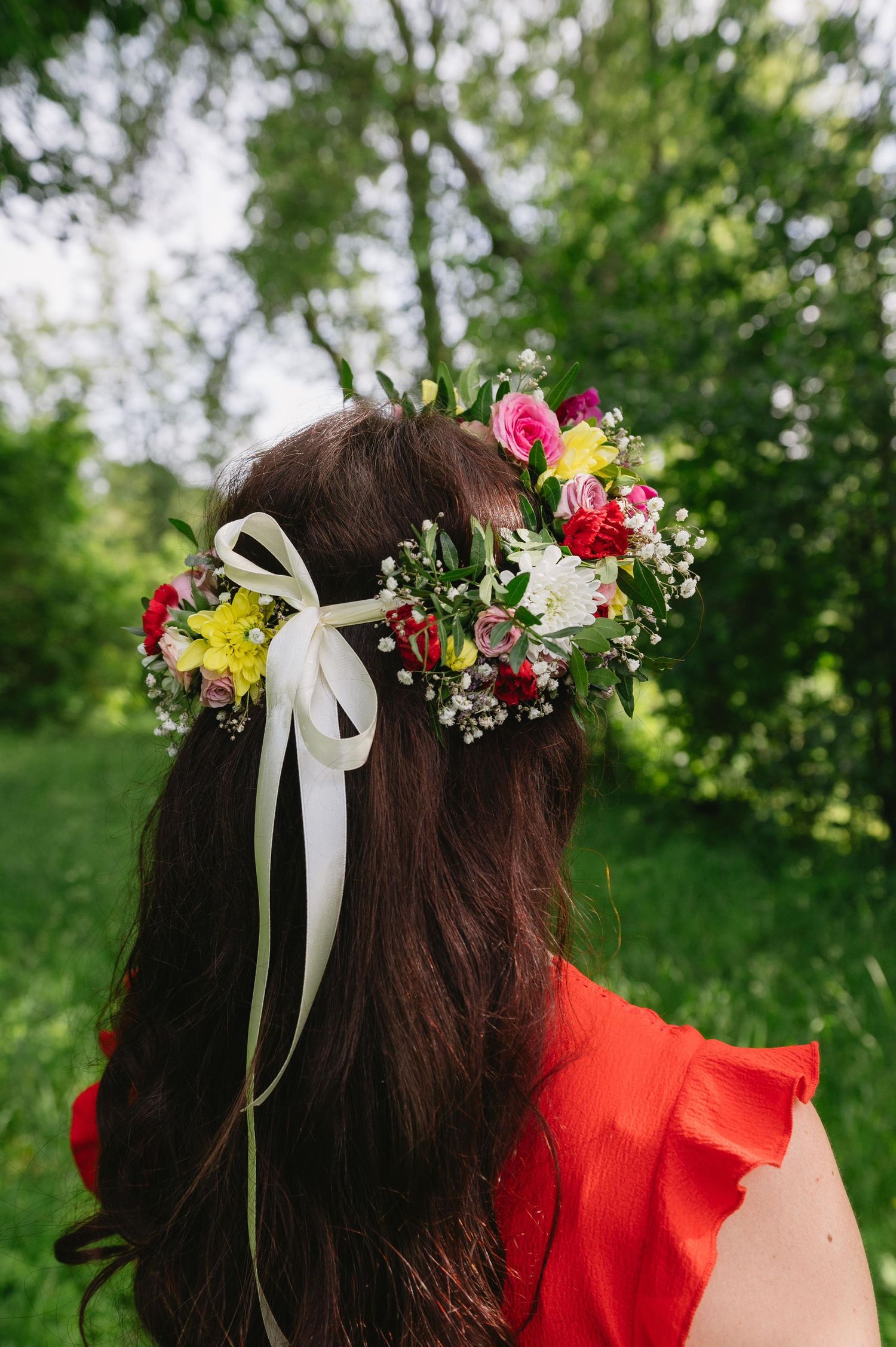 Woman wearing a colorful floral crown with ribbon bow, standing outdoors among green trees, wearing a red dress.