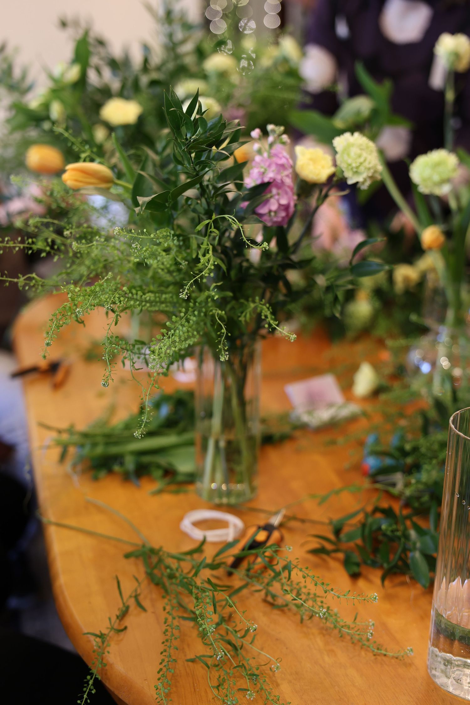 Floral arrangement workshop with green ferns and colorful flowers in glass vases on a wooden table with scissors nearby.