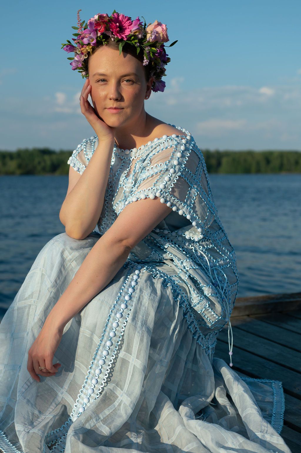 Figure in ethereal blue and white lace dress with floral crown sits on wooden dock by serene lake at sunset.