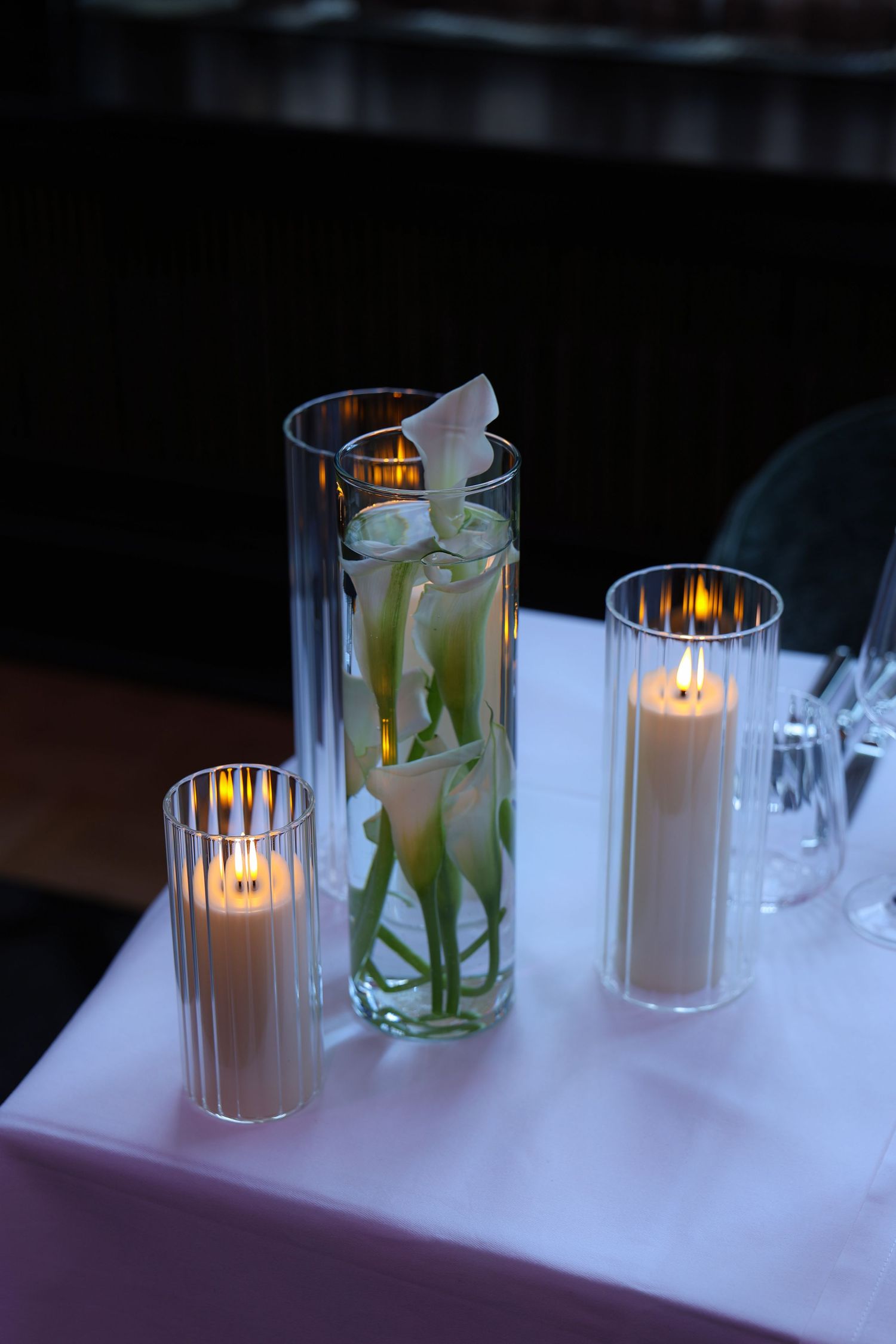 Elegant table centerpiece with calla lilies submerged in tall glass vase flanked by two lit pillar candles on white tablecloth.