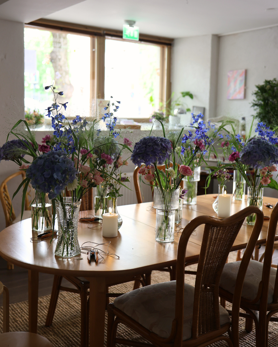 Wooden dining table with multiple glass vases of blue hydrangeas and wildflowers in a bright, airy room.