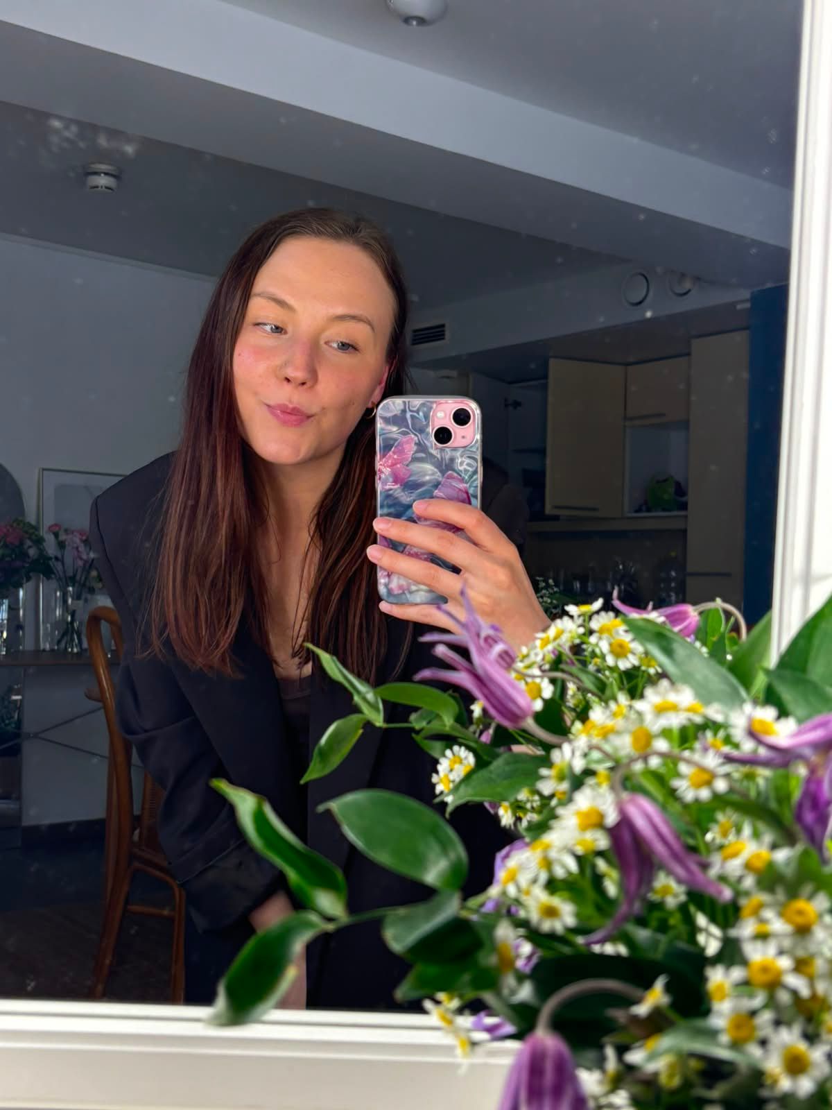 Woman taking a mirror selfie with a floral phone case, surrounded by fresh flowers and daisies in a bright room.
