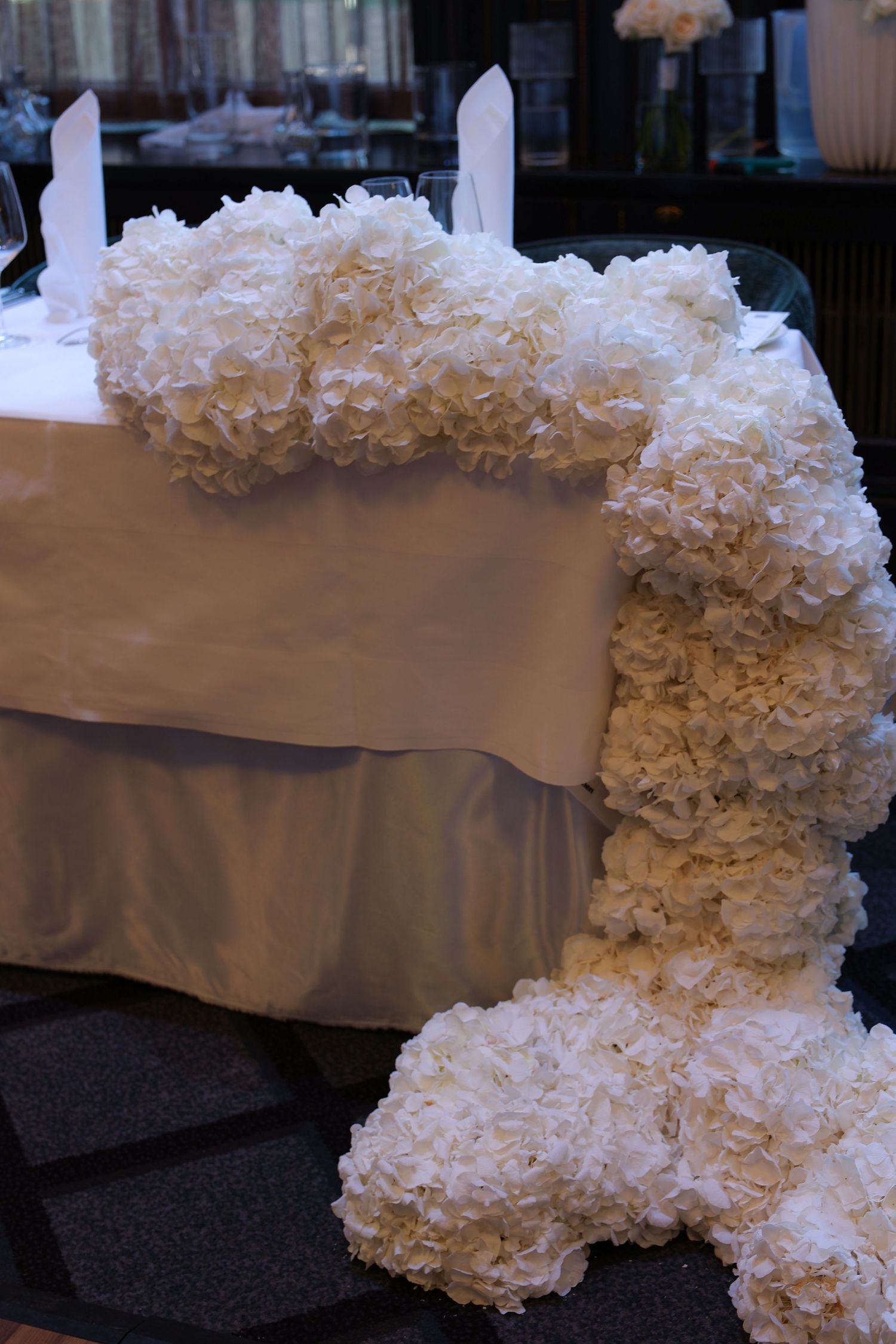 Cascading white ruffled fabric or floral decorations arranged along a table edge with a taupe backdrop.