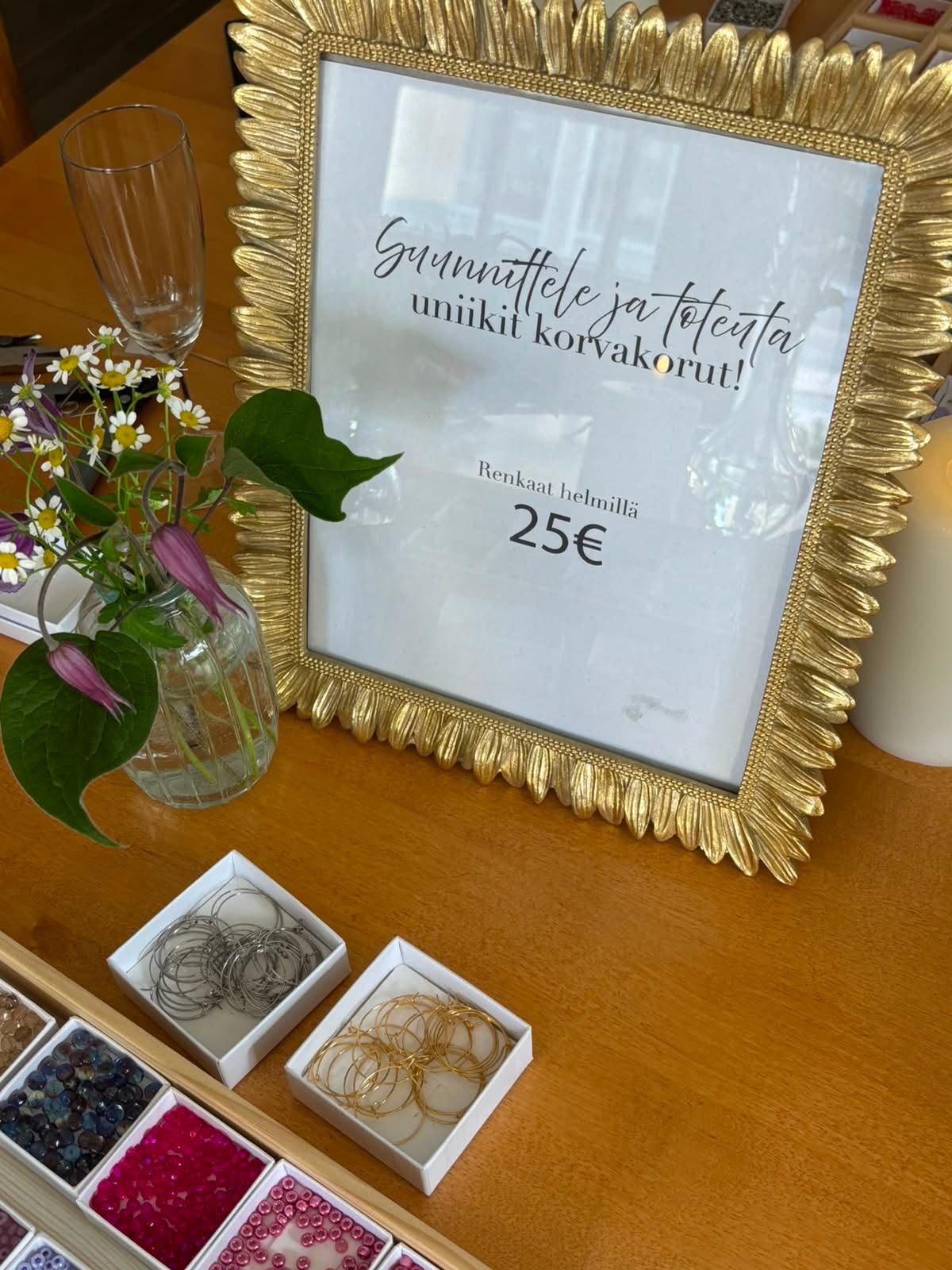 Gold framed sign advertising unique handmade earrings for 25 euros, displayed with beads and wire on a wooden table.