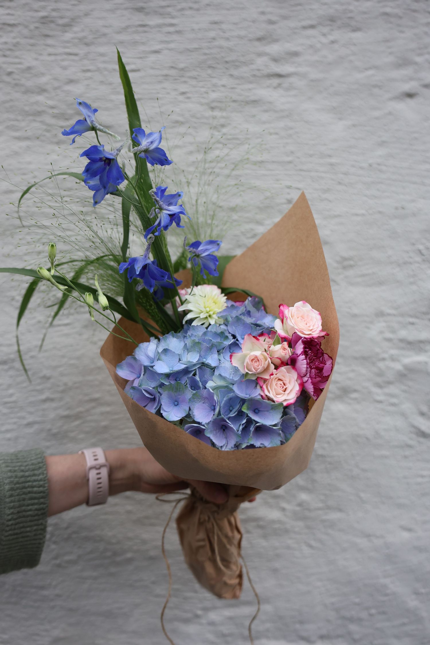 Hand holding a bouquet of blue hydrangeas, pink roses, and delphinium wrapped in kraft paper.
