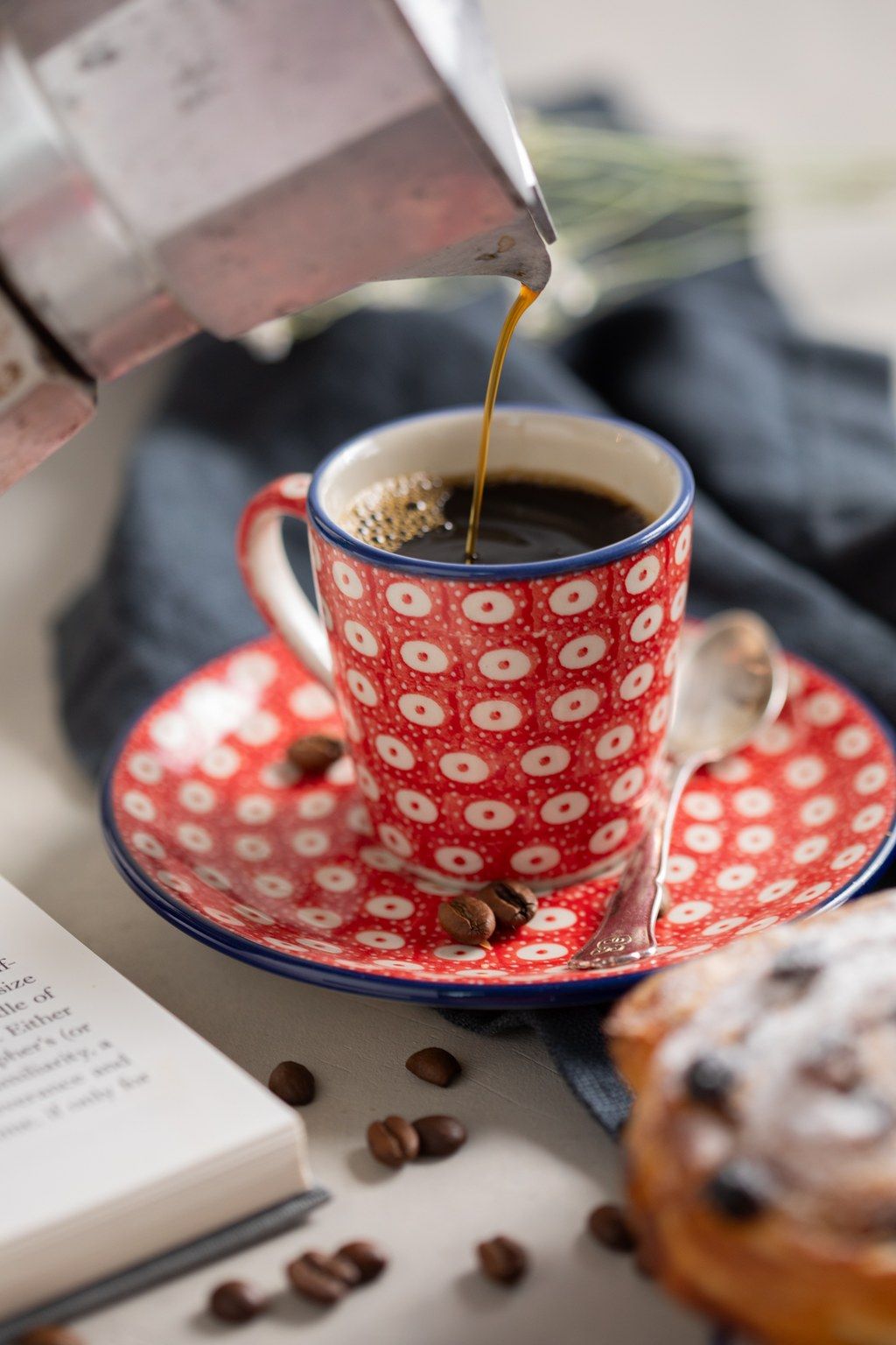 Coffee being poured into a red and white patterned teacup with coffee beans and pastry on the side.