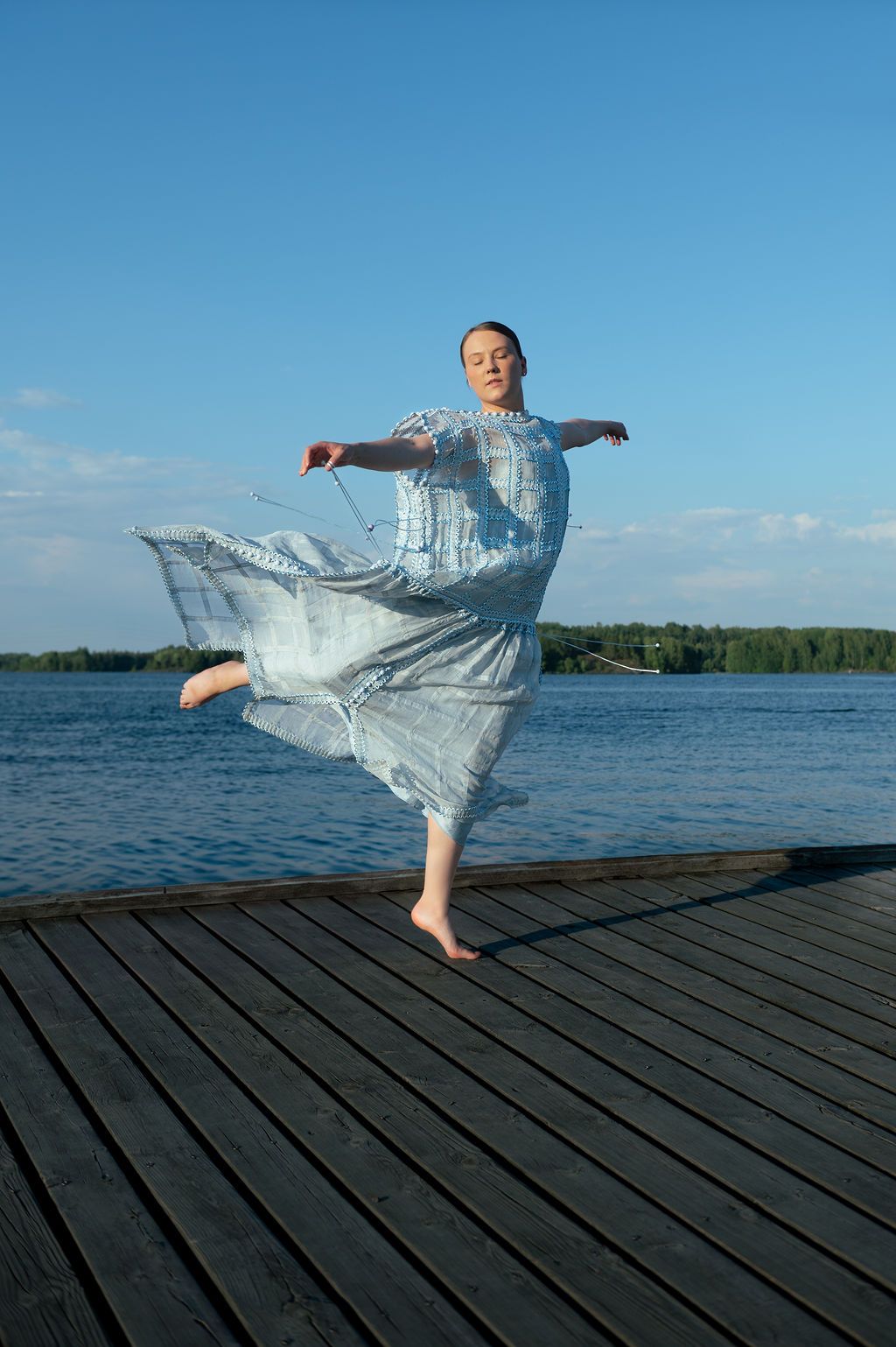 A figure in a flowing silver dress performs an elegant dance sequence on a wooden dock by the water.