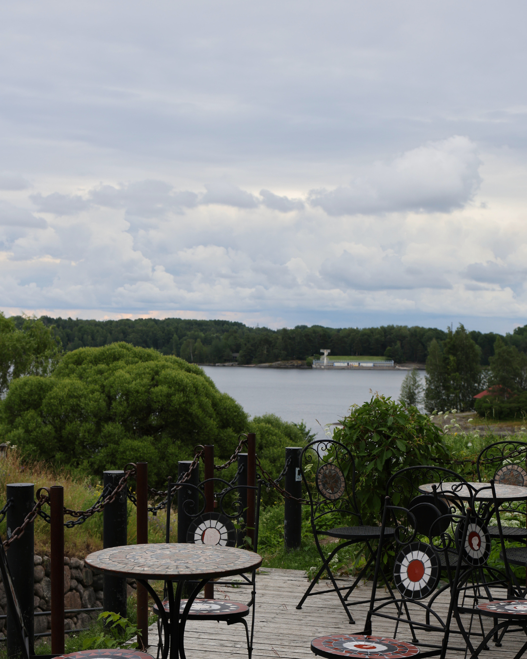 Outdoor cafe terrace with mosaic tables and iron chairs overlooking a serene lake surrounded by lush green trees under cloudy skies.