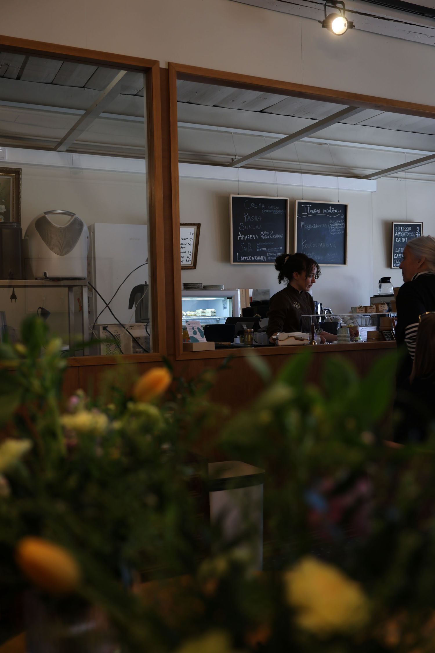 Interior view of a modern cafe with wooden framed windows and blurred greenery in the foreground.