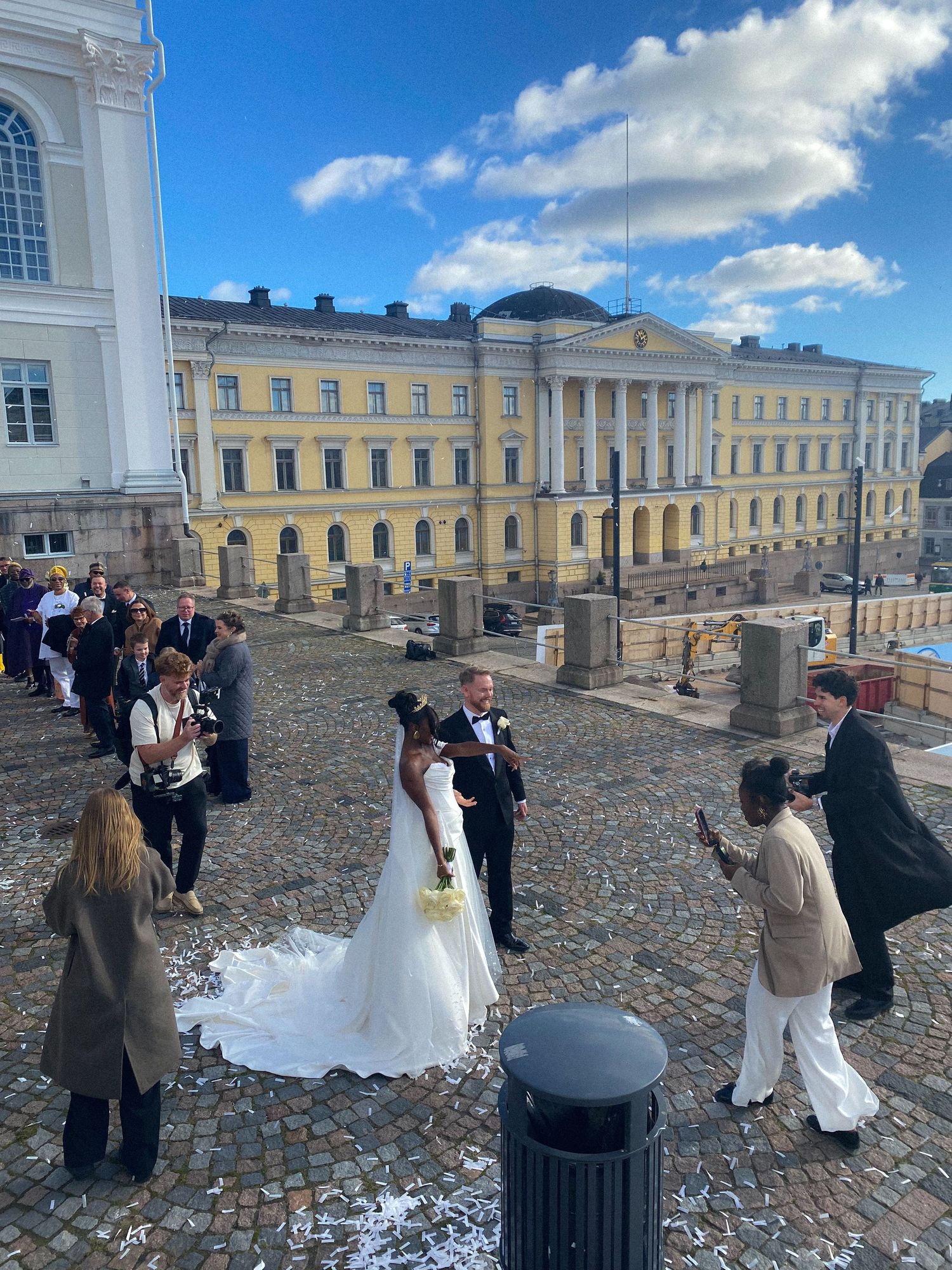 Wedding couple posing on Helsinki Senate Square steps with yellow government building backdrop on sunny day.