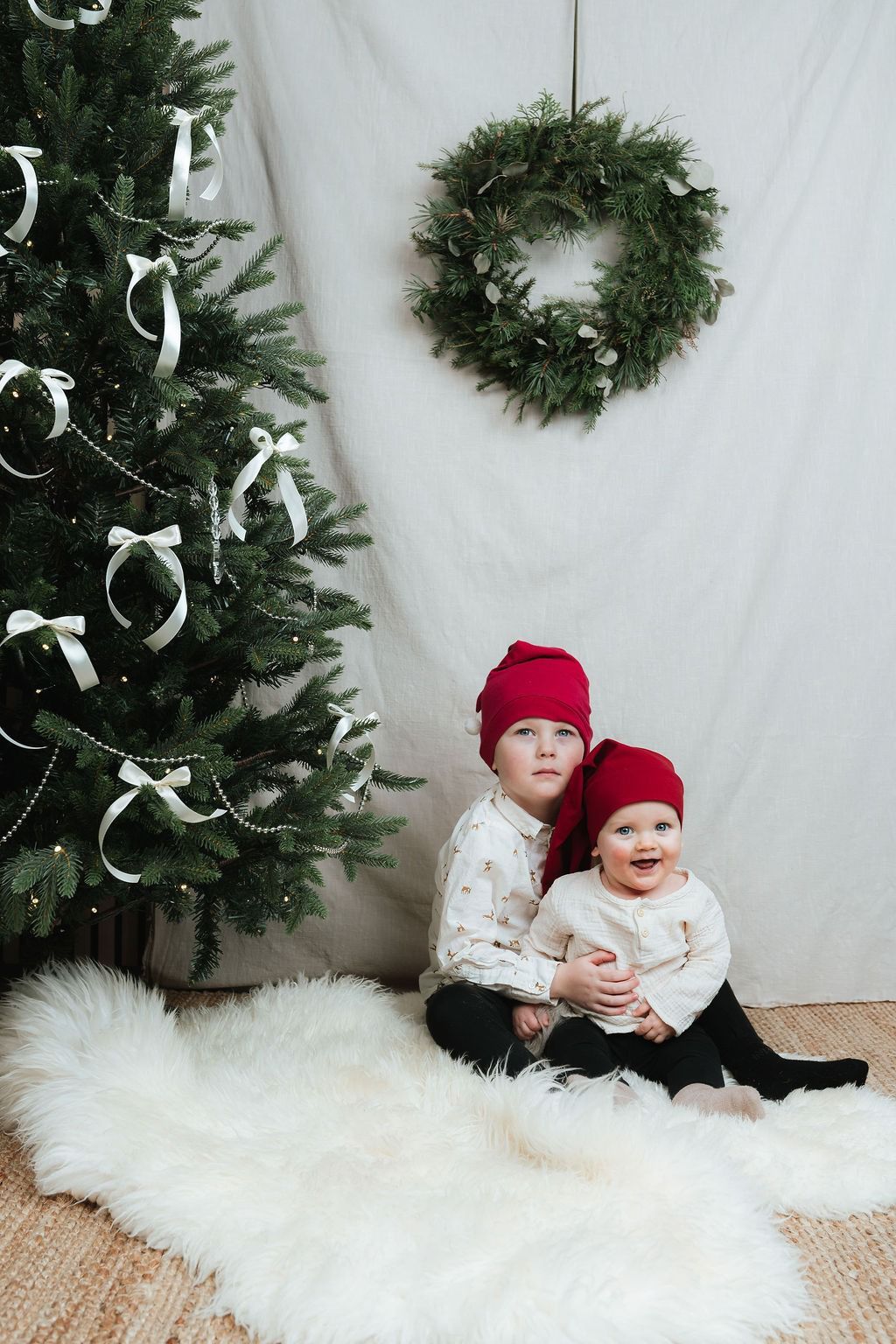 Progressive series of baby in red hat and white dress sitting on fluffy white rug by Christmas tree and wreath.