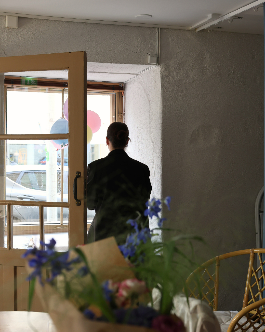 Person standing silhouetted by a bright window in a minimalist cafe with blue flowers in the foreground.