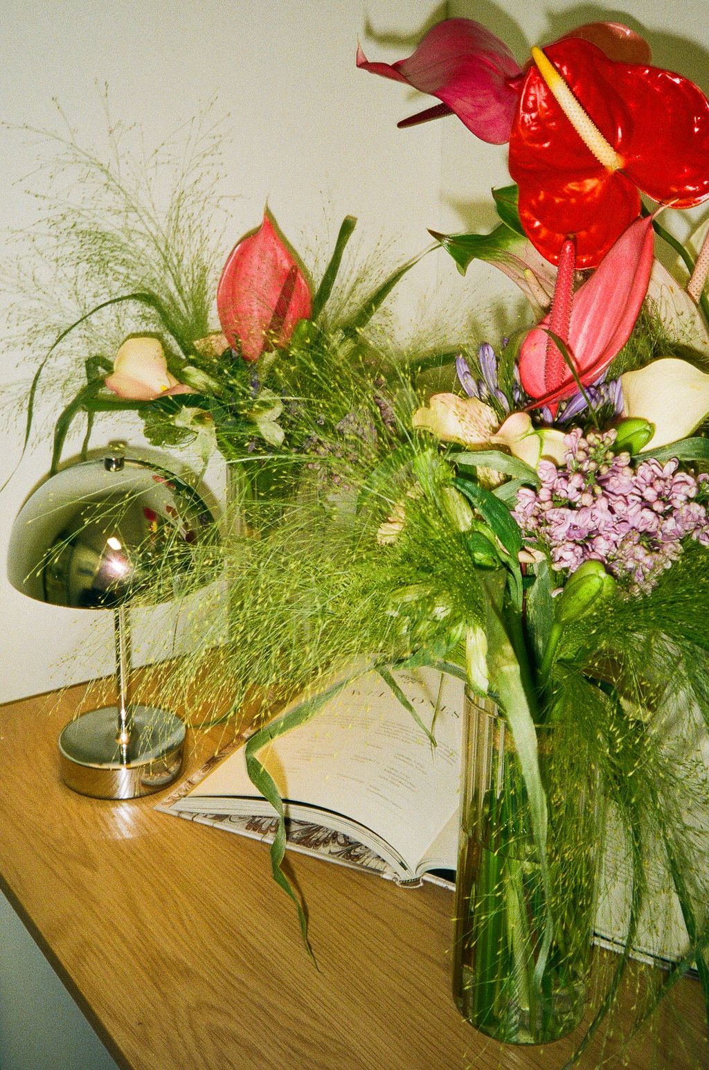 Red anthuriums and pink flowers in a glass vase beside a chrome lamp on a wooden desk.