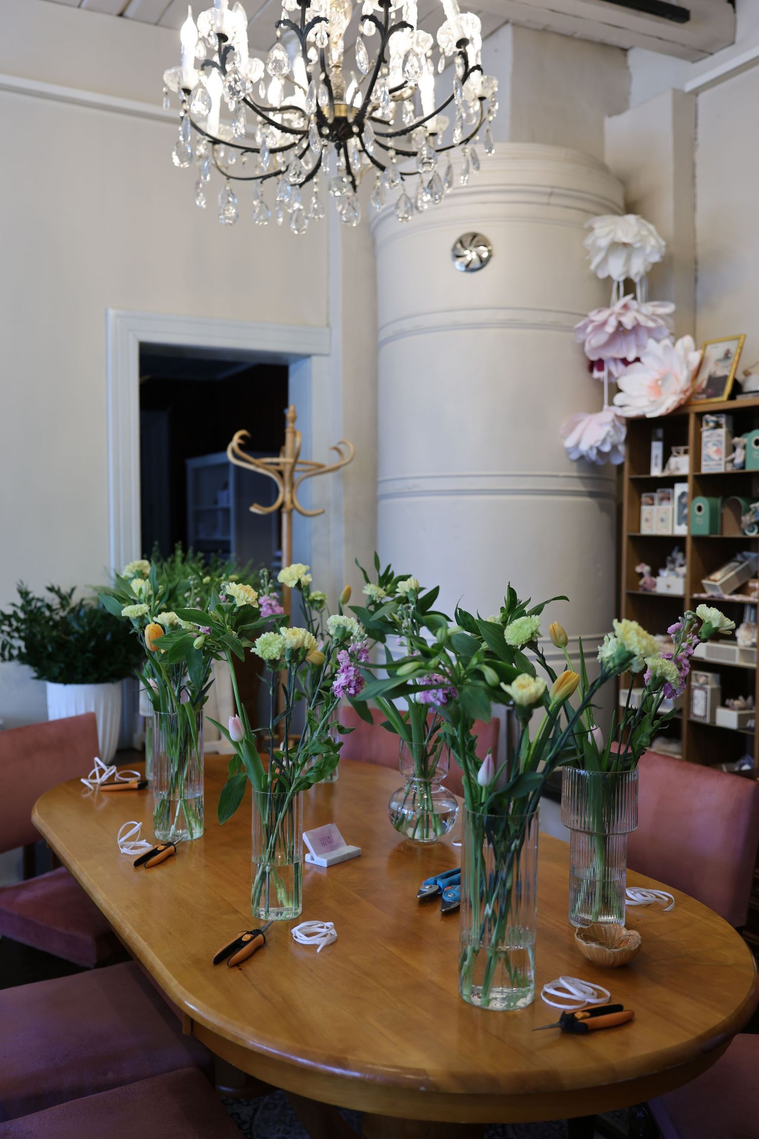 A wooden table with purple flowers in glass vases sits beneath an elegant crystal chandelier in a bright interior space.
