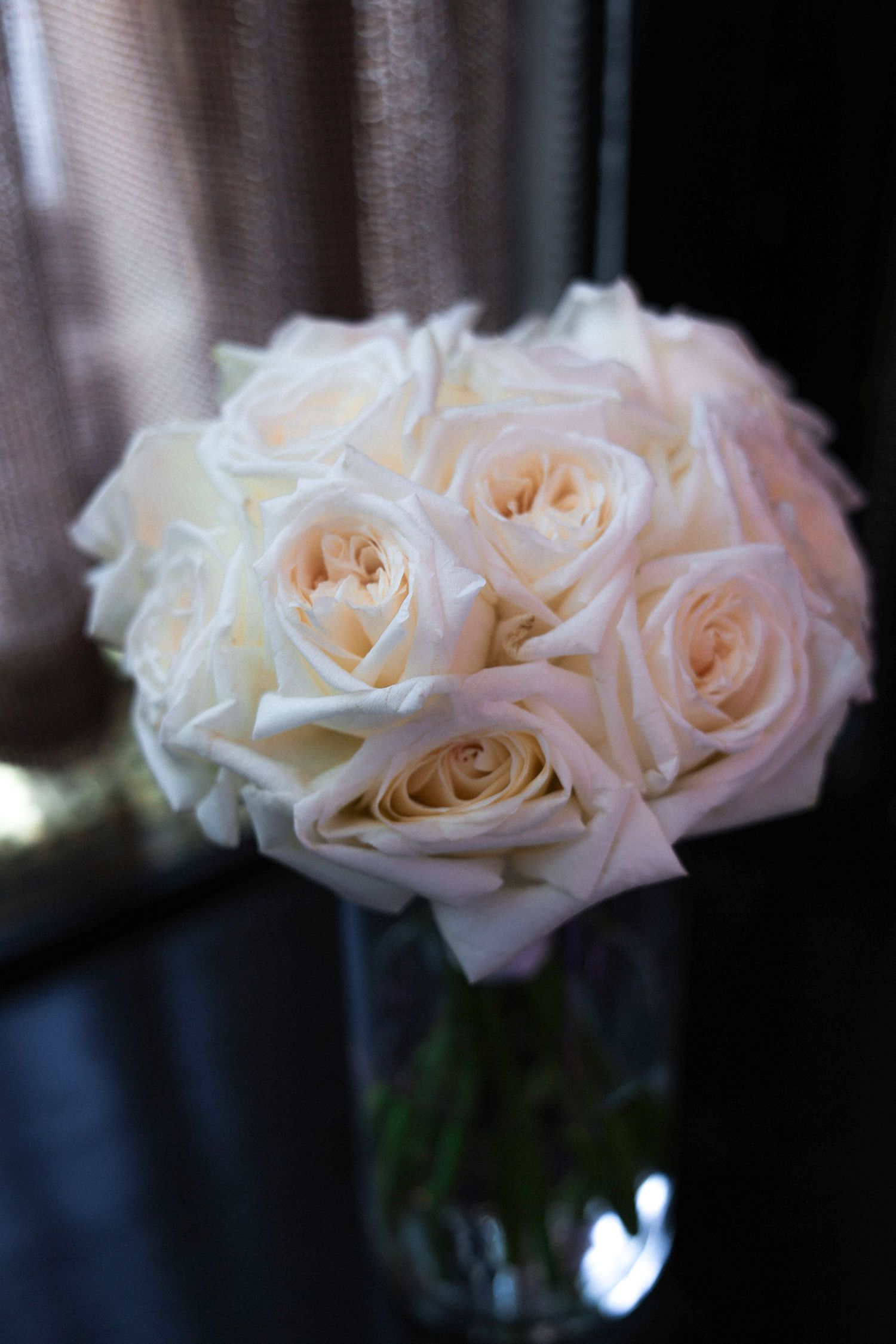 Delicate cream-colored roses with soft peach undertones arranged in a simple glass vase against a dark background.