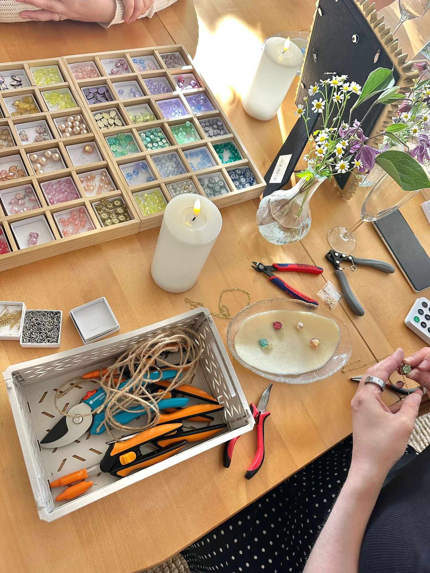 Jewelry making workspace with colorful bead organizer, pliers, wire, tools, candle, and floral arrangement on wooden table.