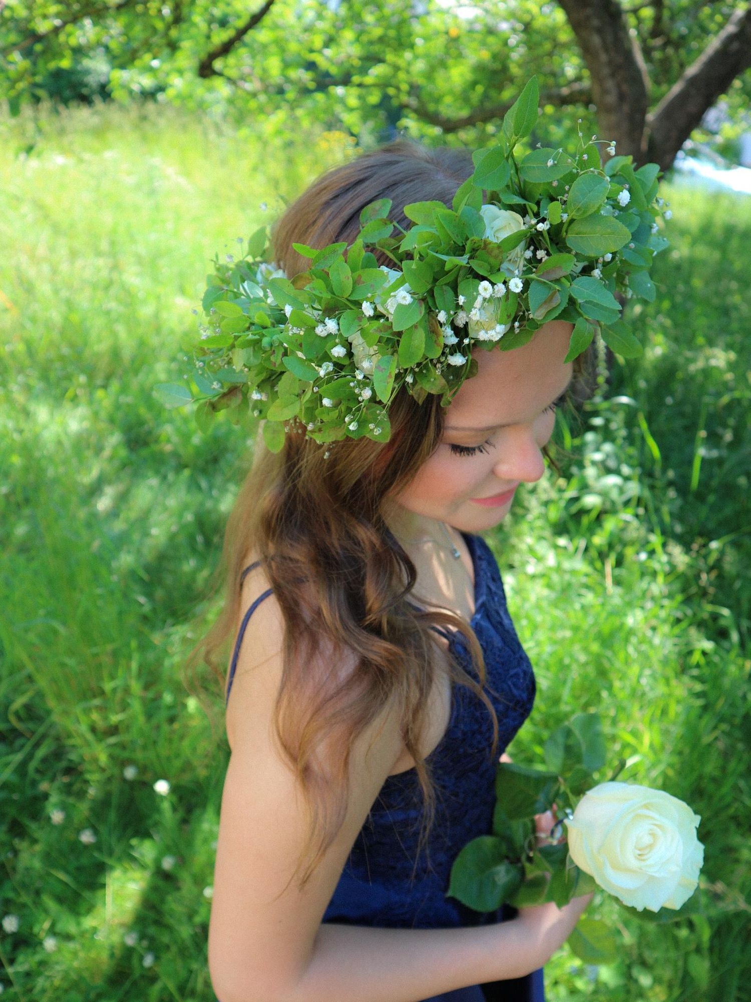 A serene nature scene shows a person in a navy dress adorned with a delicate floral crown among lush green foliage.
