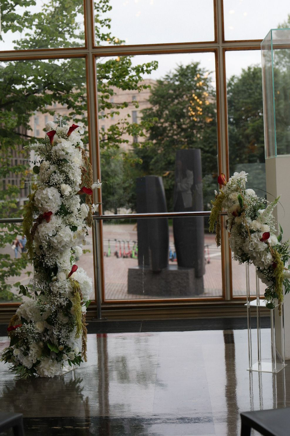 Elegant floral arrangements with white blooms and red roses frame a large window overlooking an outdoor sculpture garden.