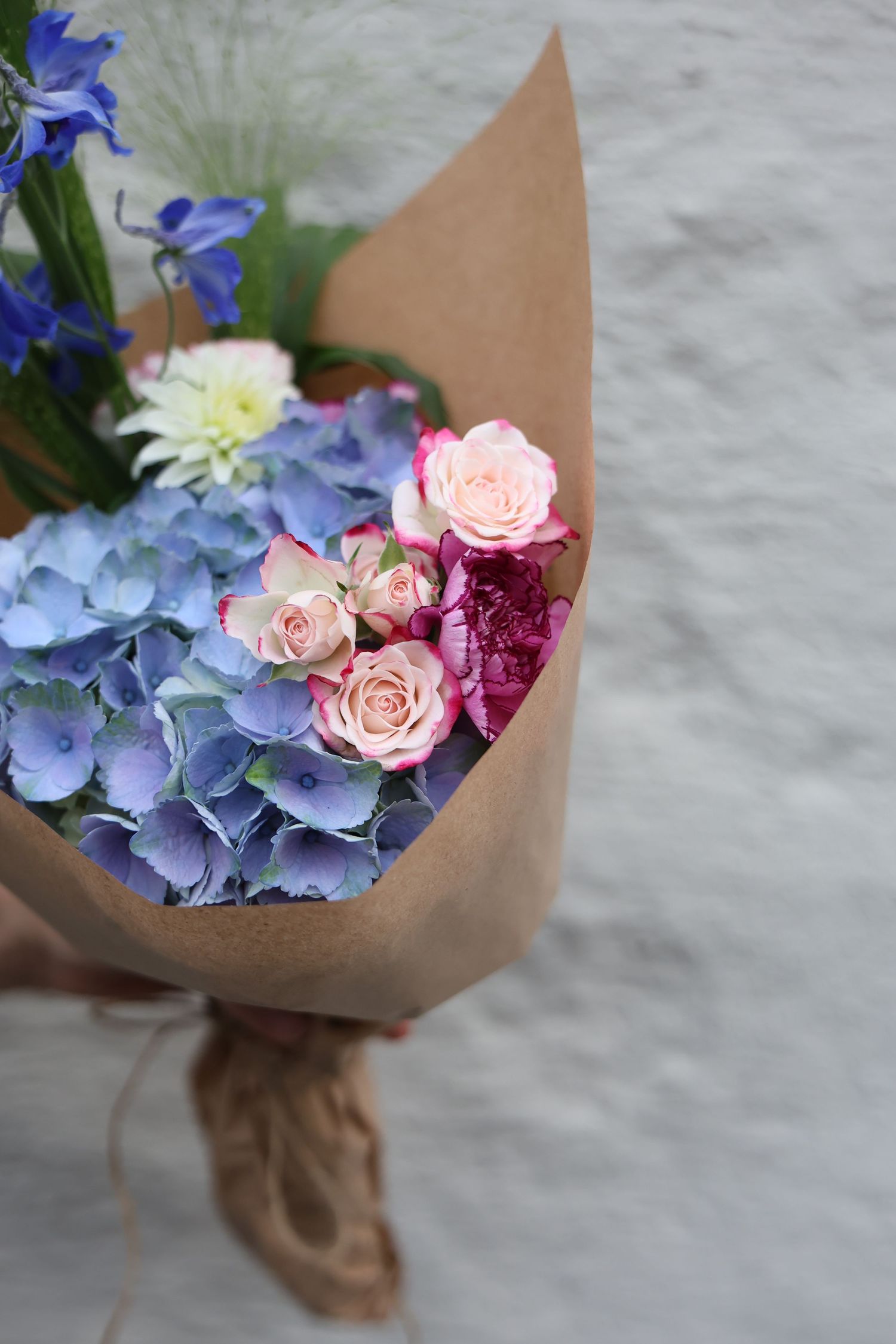 Hand-held bouquet of blue hydrangeas, pink roses, and mixed flowers wrapped in kraft paper.