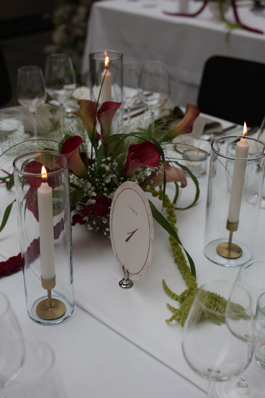 Wedding reception table with lit candles in glass holders, red calla lilies, and an oval table number card.