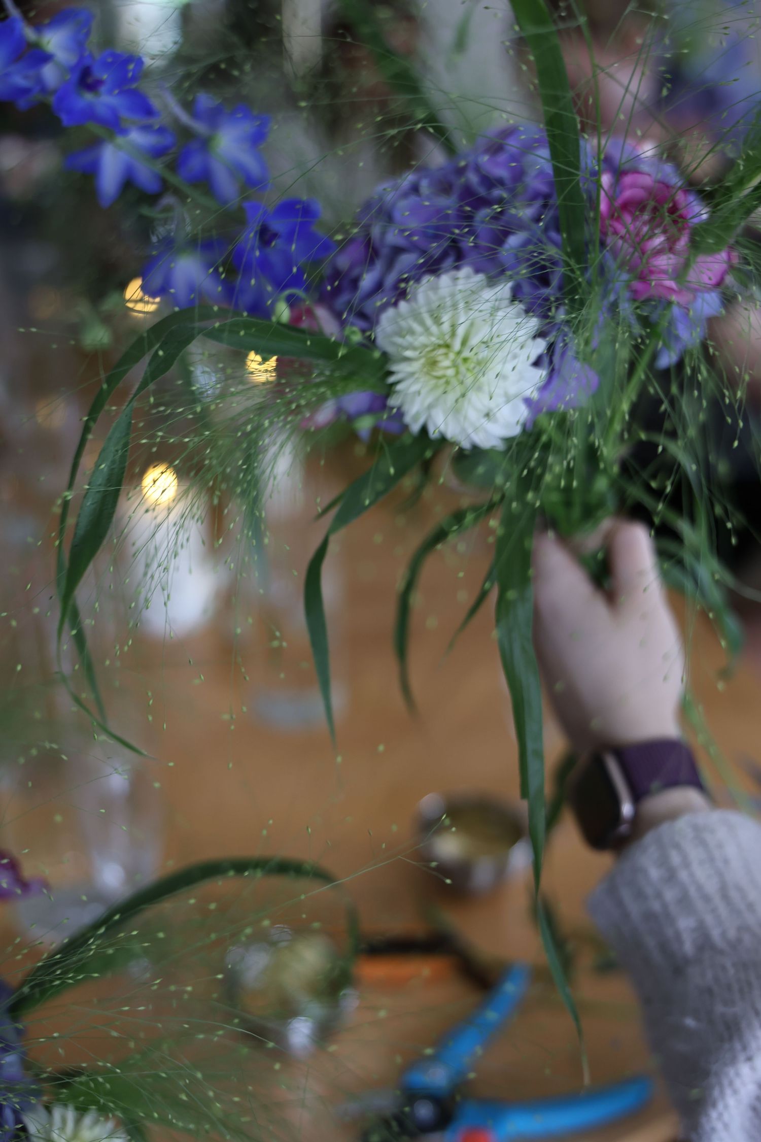 Hand arranging purple, blue, and white flowers with green foliage, viewed through a rain-speckled window.