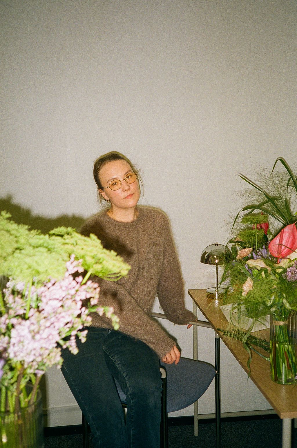 Woman with glasses in brown sweater sits among colorful flower arrangements on a table in a bright room.