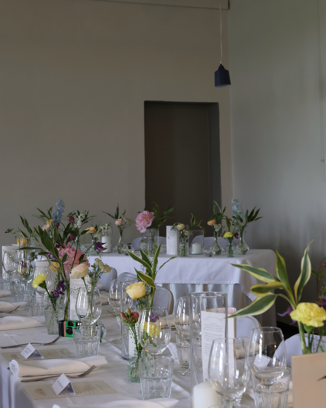 Elegant wedding reception table set with white linens, glass vases of pastel flowers, and crystal glassware in a minimalist venue.