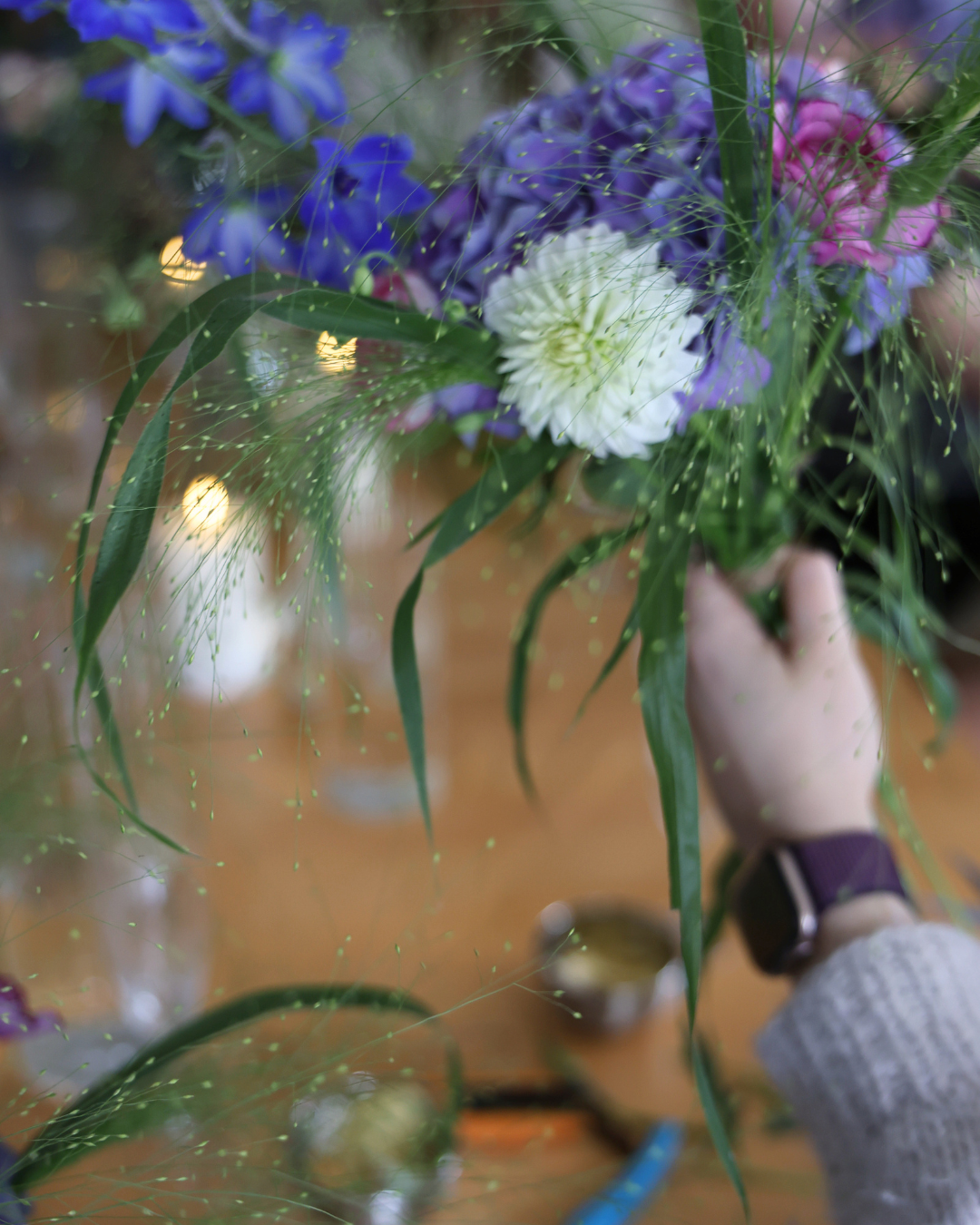 Florist arranging blue, purple, and white flowers with green foliage over a reflective surface.