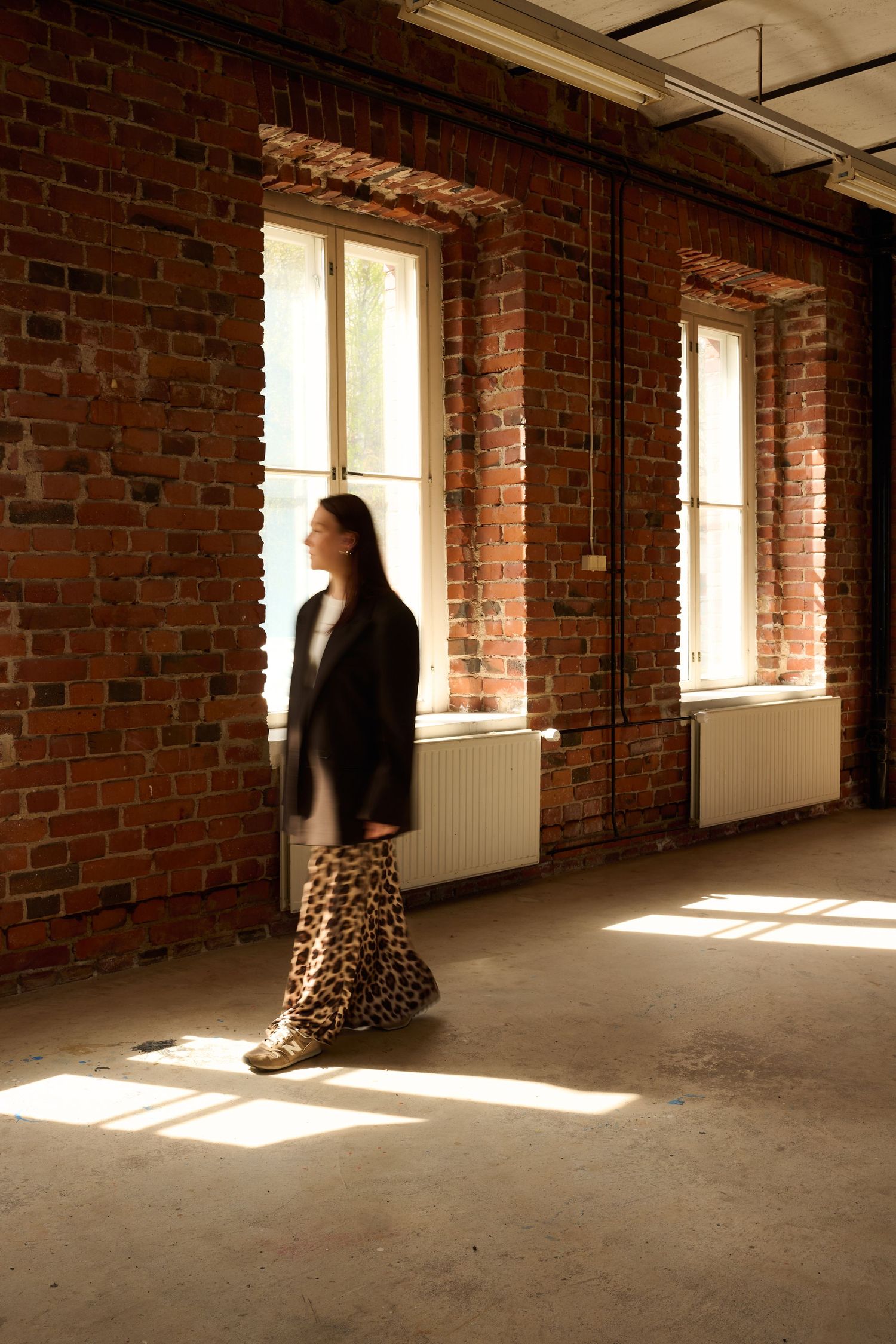Woman in leopard skirt and black blazer walking in sunlit brick room with large windows casting geometric shadows.