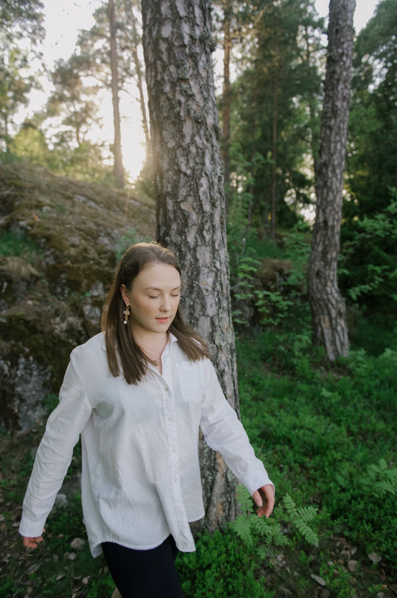 Person in white button-down shirt stands contemplatively among tall pine trees in a peaceful forest setting.