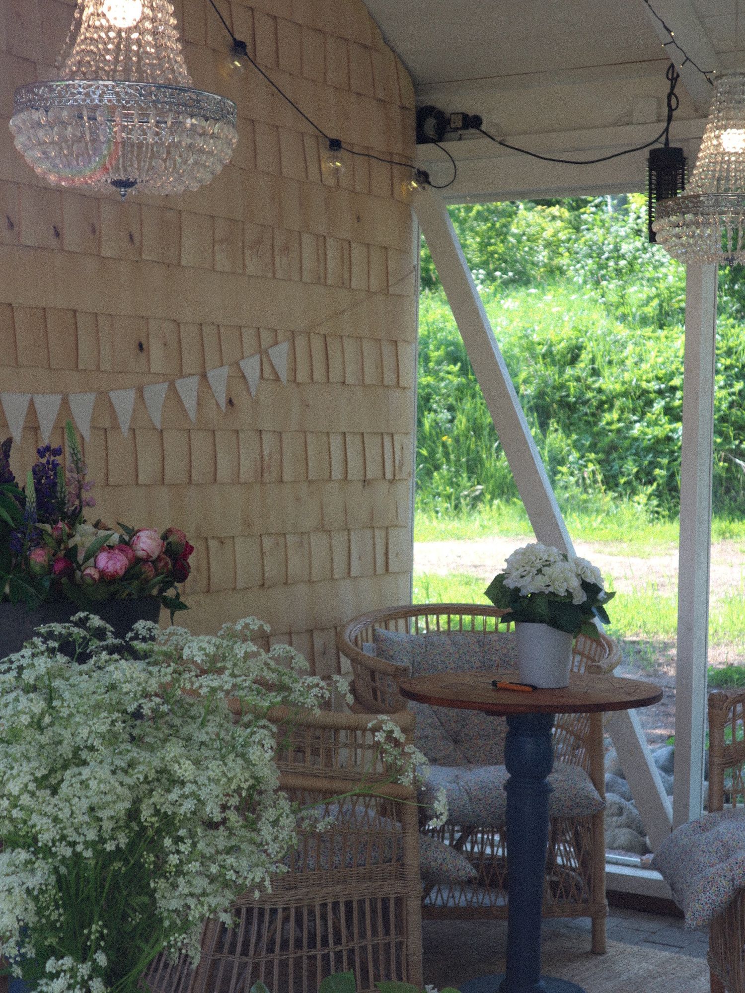 Crystal chandeliers and delicate white bunting decorate a rustic porch with wicker furniture and fresh floral arrangements.
