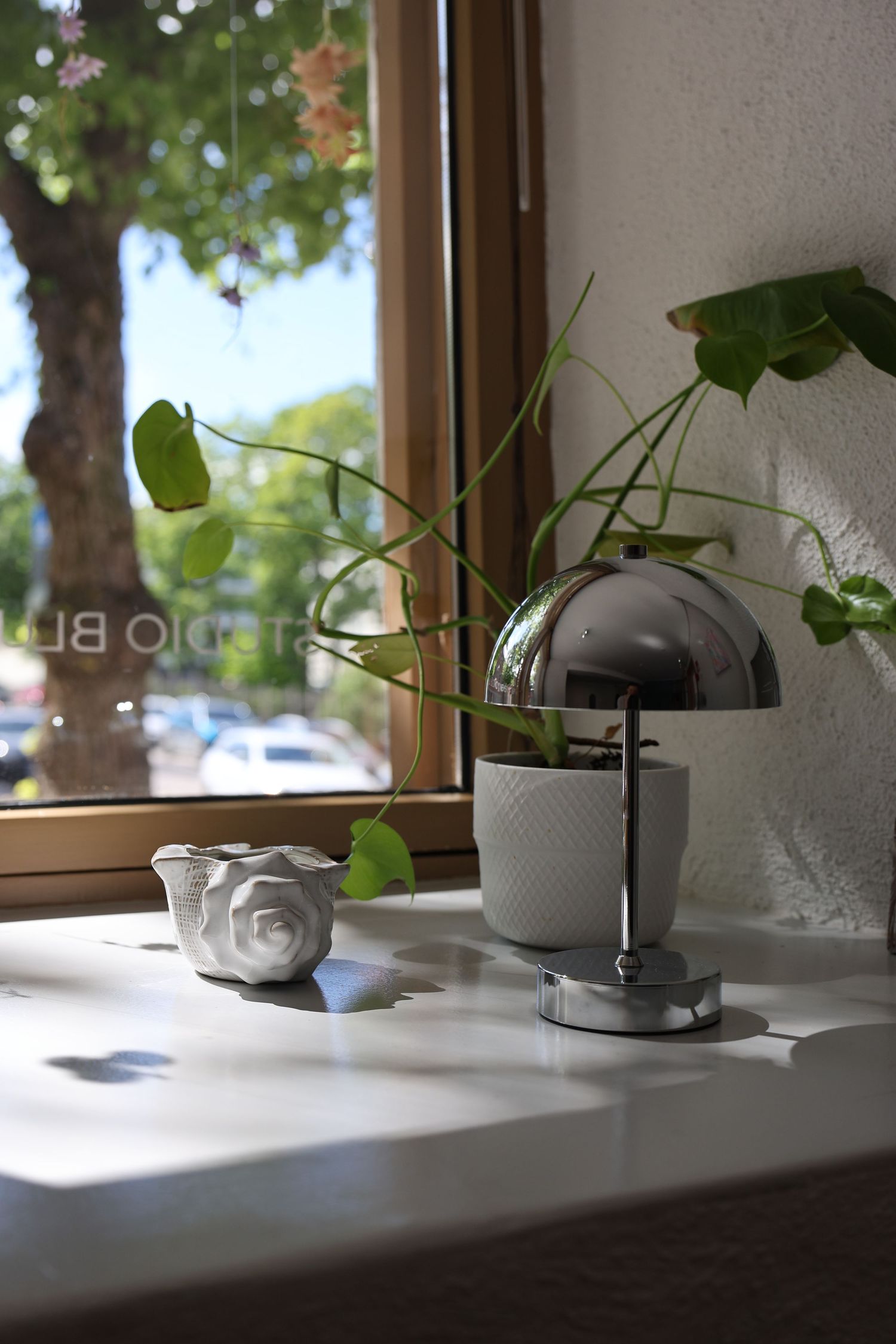 Chrome dome lamp and green potted plant on white windowsill with wooden frame window and tree view outside.