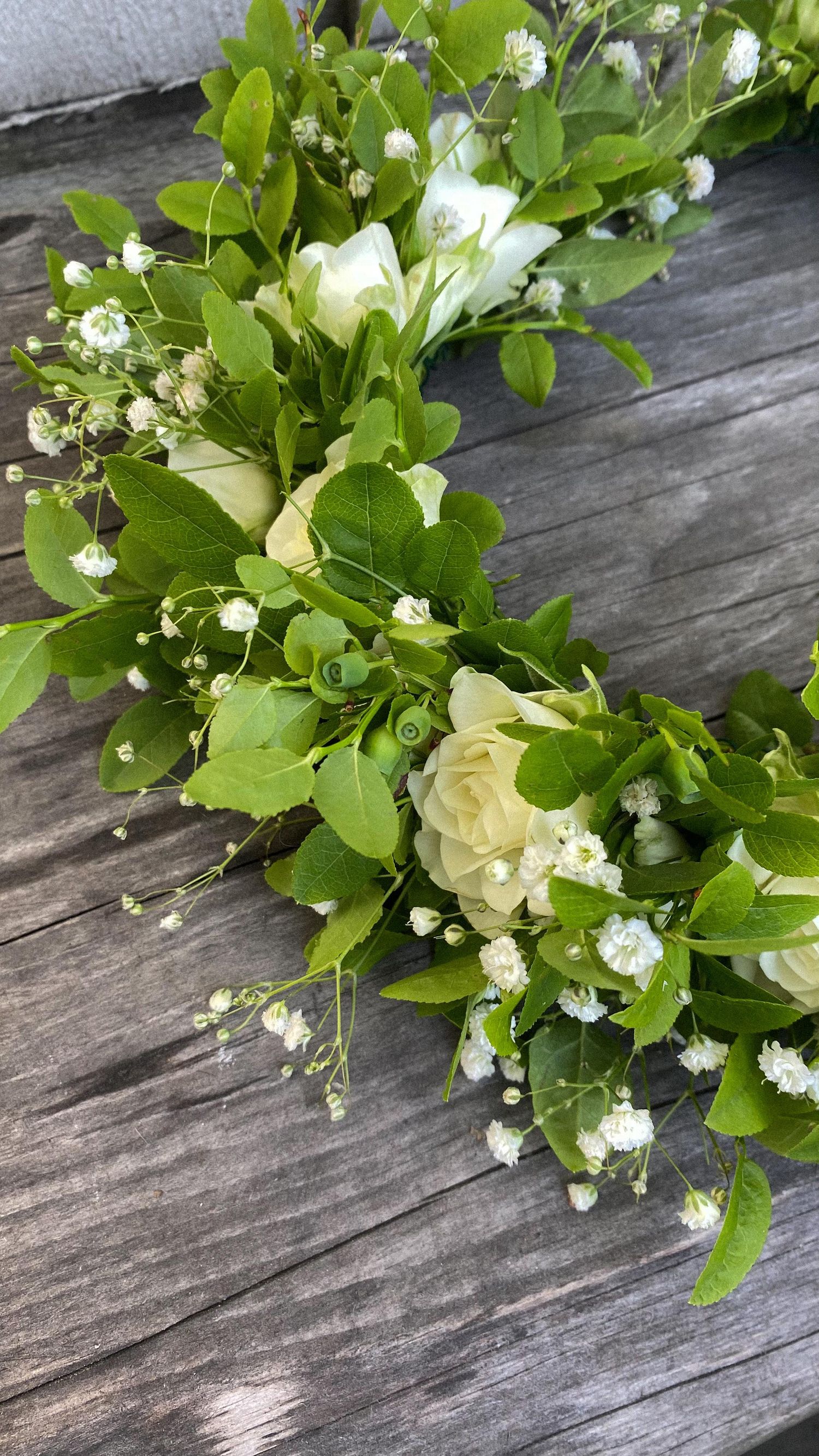A delicate floral arrangement with white roses and baby's breath nestled among fresh green leaves on a weathered wood surface.