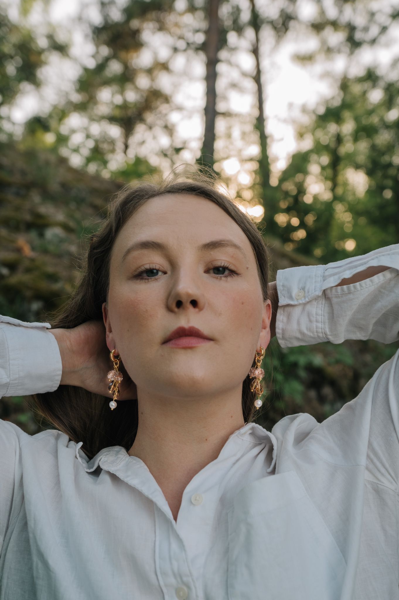 Young woman in white shirt with gold earrings poses outdoors in forest setting with hands behind her head.