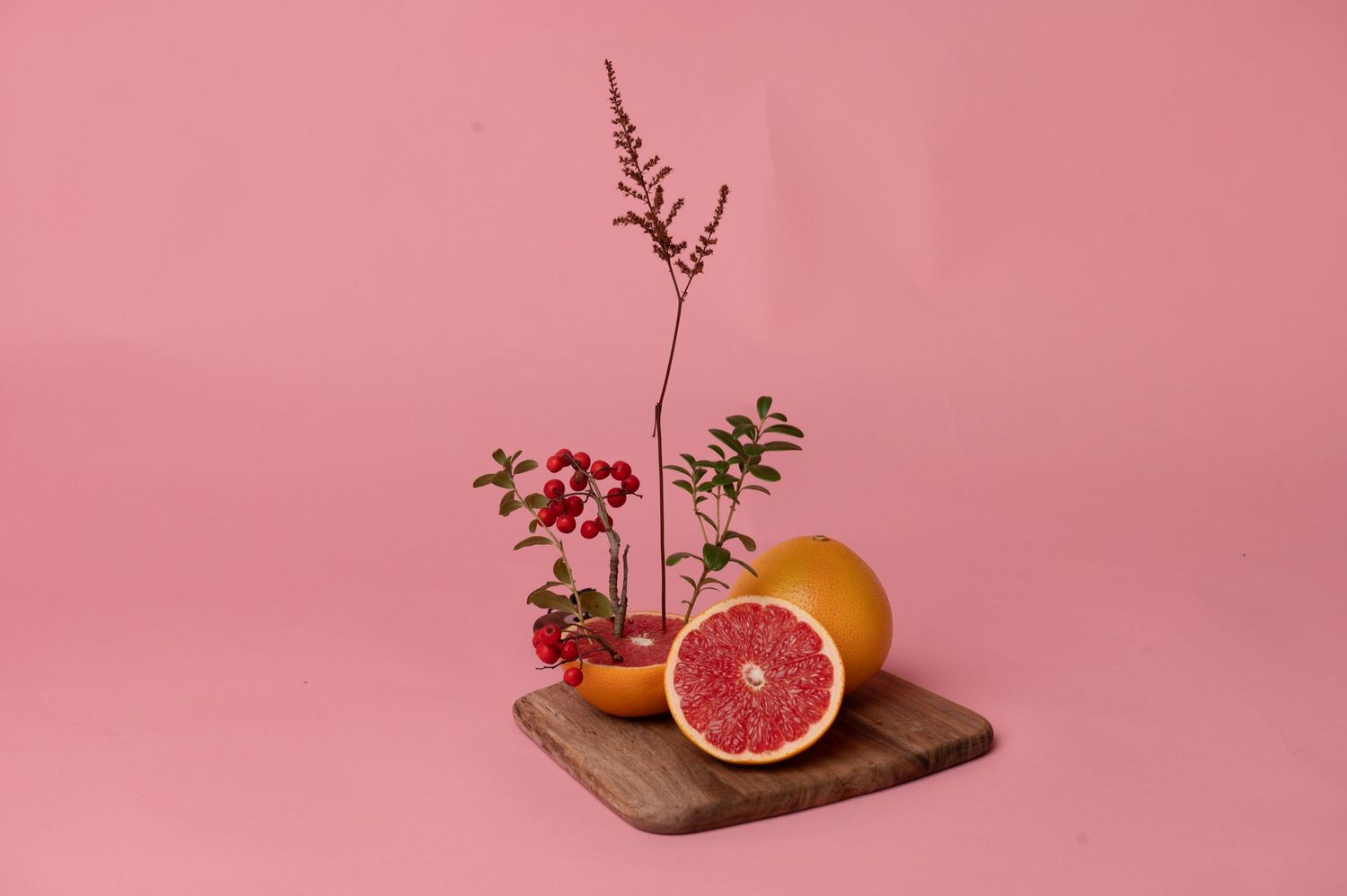 Halved grapefruit and whole orange on wooden board with red berries and branches against pink background.