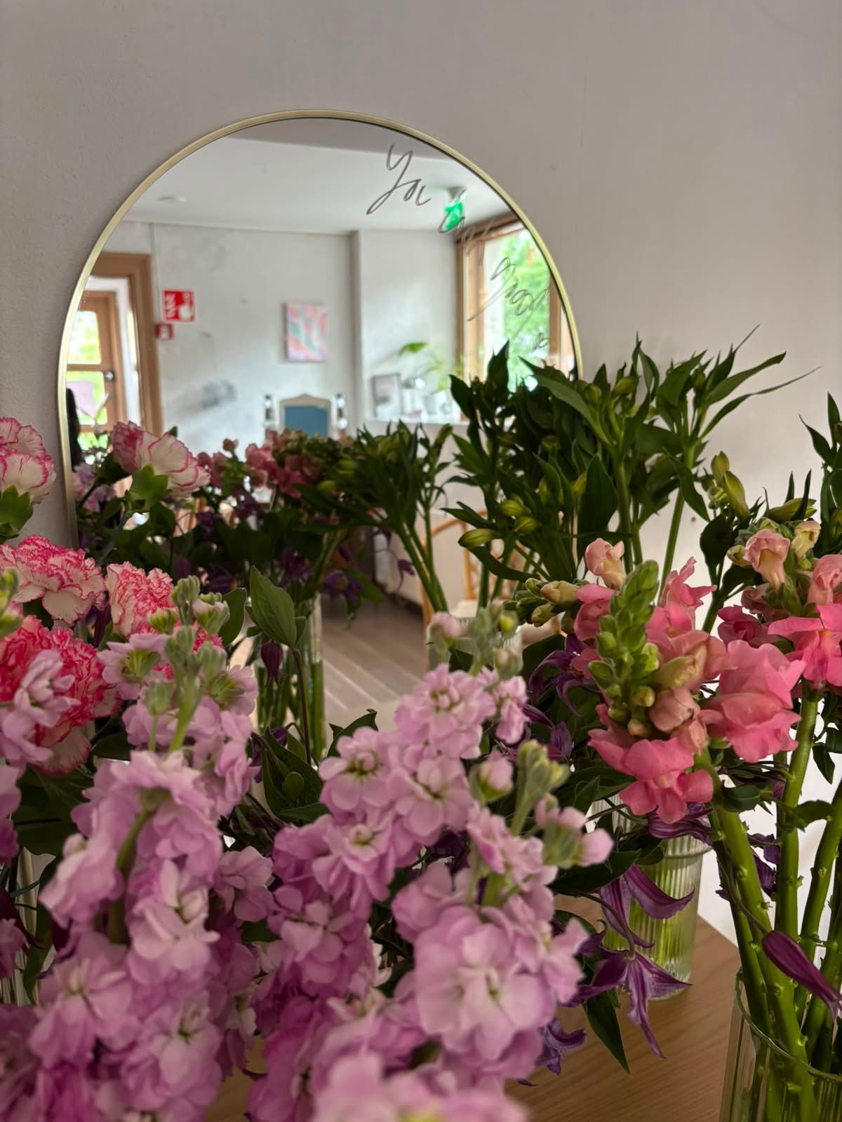 Round gold mirror surrounded by lush pink blooms including snapdragons and stock flowers in a bright floral studio.