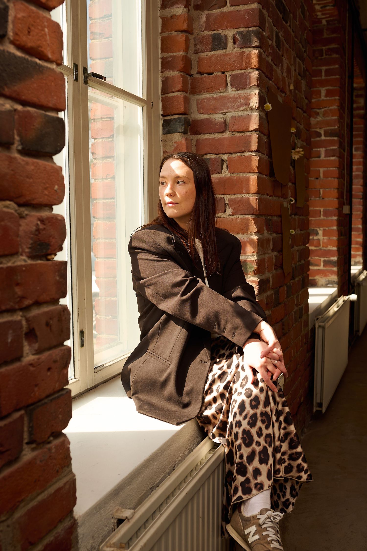 Woman in oversized blazer and leopard print skirt sits on windowsill beside brick wall, gazing thoughtfully.