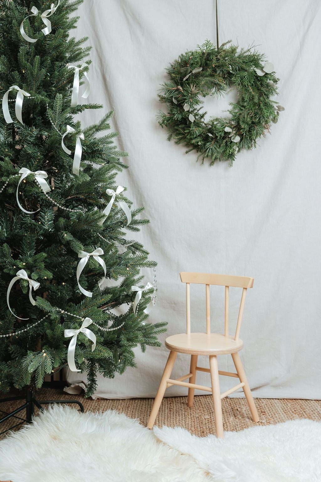 Minimalist Christmas decor with decorated green tree, wooden chair, and wreath on white backdrop.