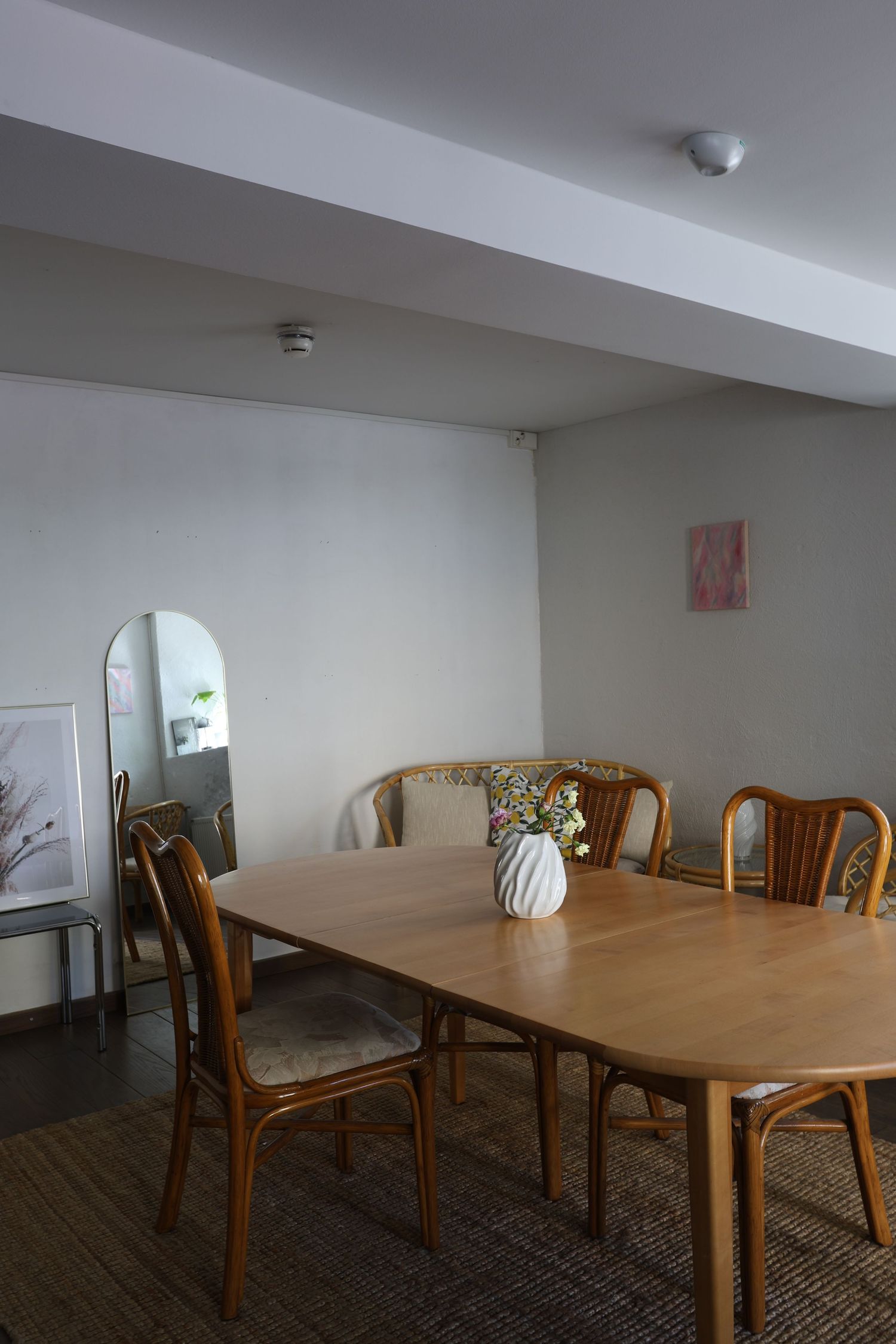 Minimalist dining room with wooden table, rattan chairs, white vase, arched mirror, and soft gray walls on a jute rug.