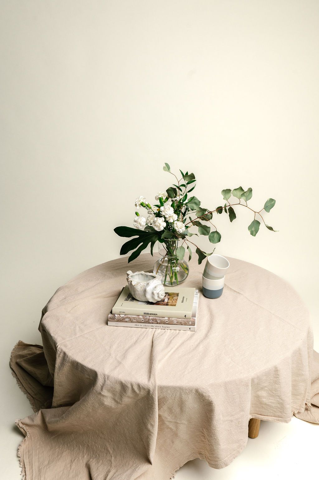 A minimalist table setting with white flowers in a glass vase, stacked books, and a beige linen tablecloth.