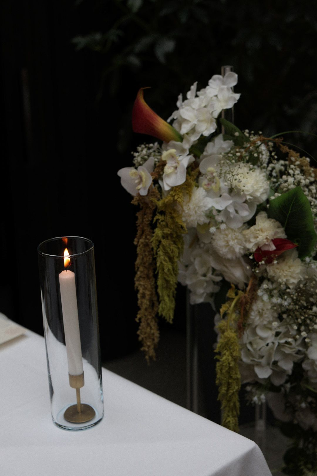 Elegant wedding table with lit white candle in glass vase and white floral arrangement with hydrangeas and calla lilies.