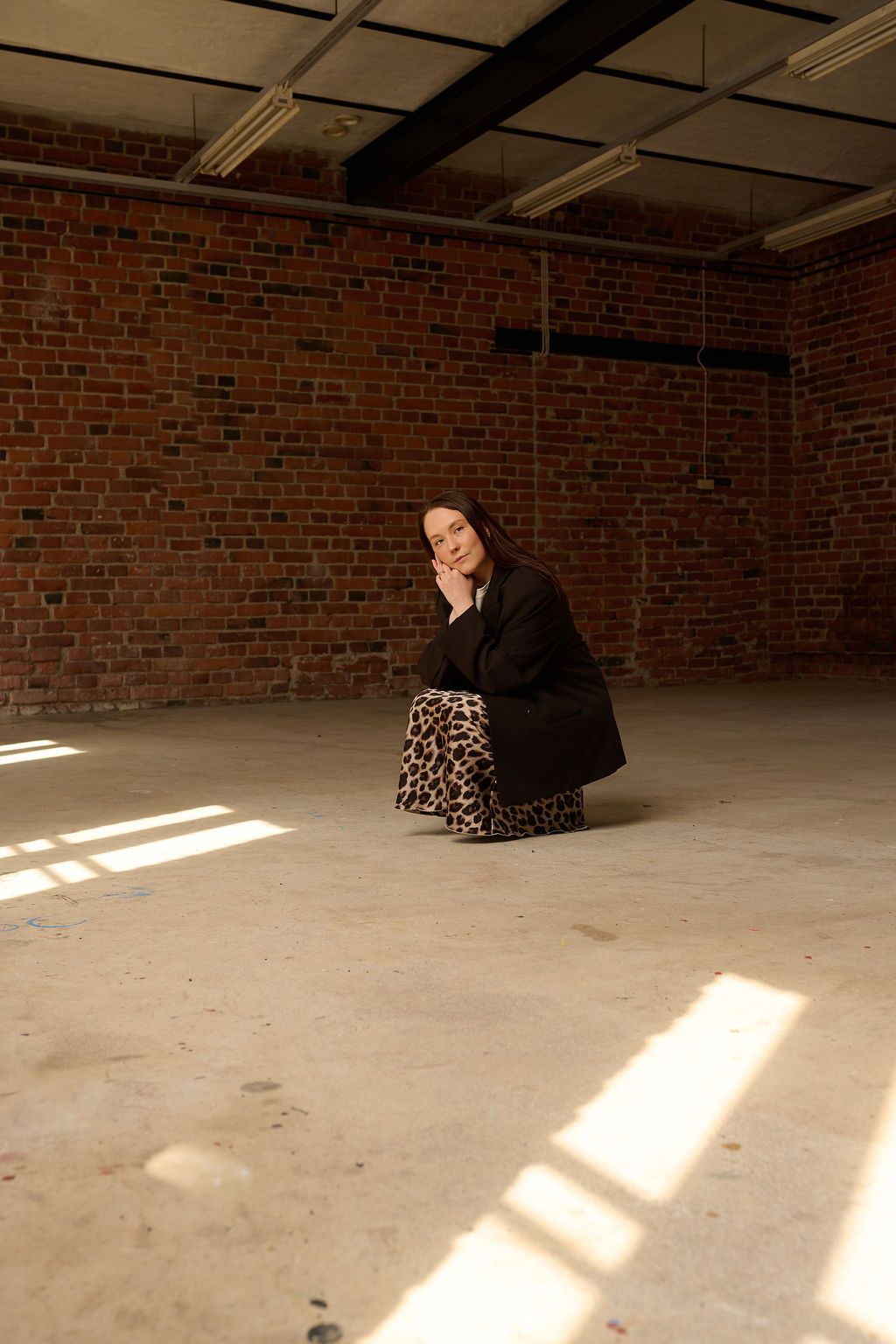 Woman in black jacket and leopard skirt sitting on concrete floor against red brick wall in sunlight.