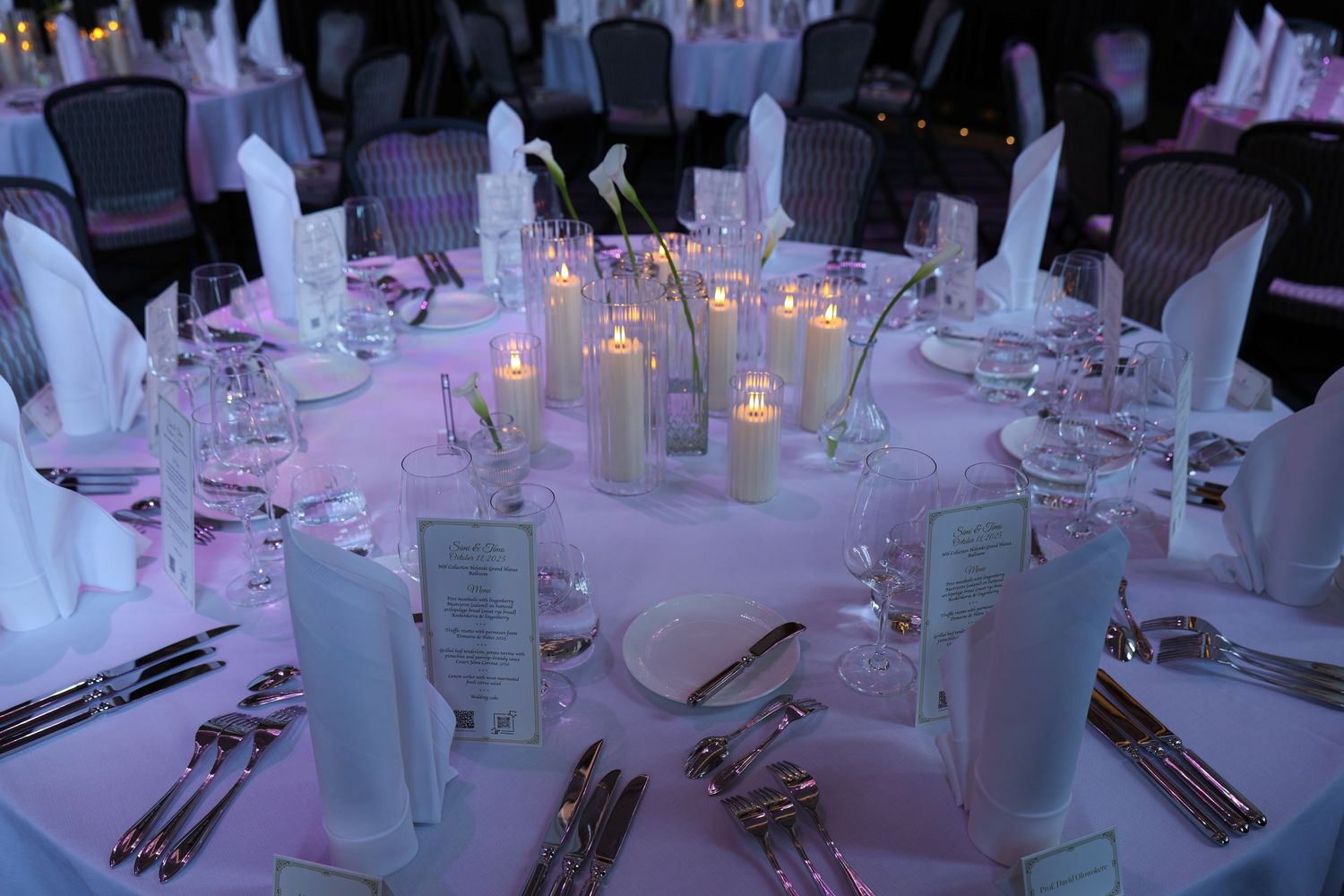 Elegant wedding reception tables decorated with white linens, glowing candles, and formal place settings in a dimly lit venue.