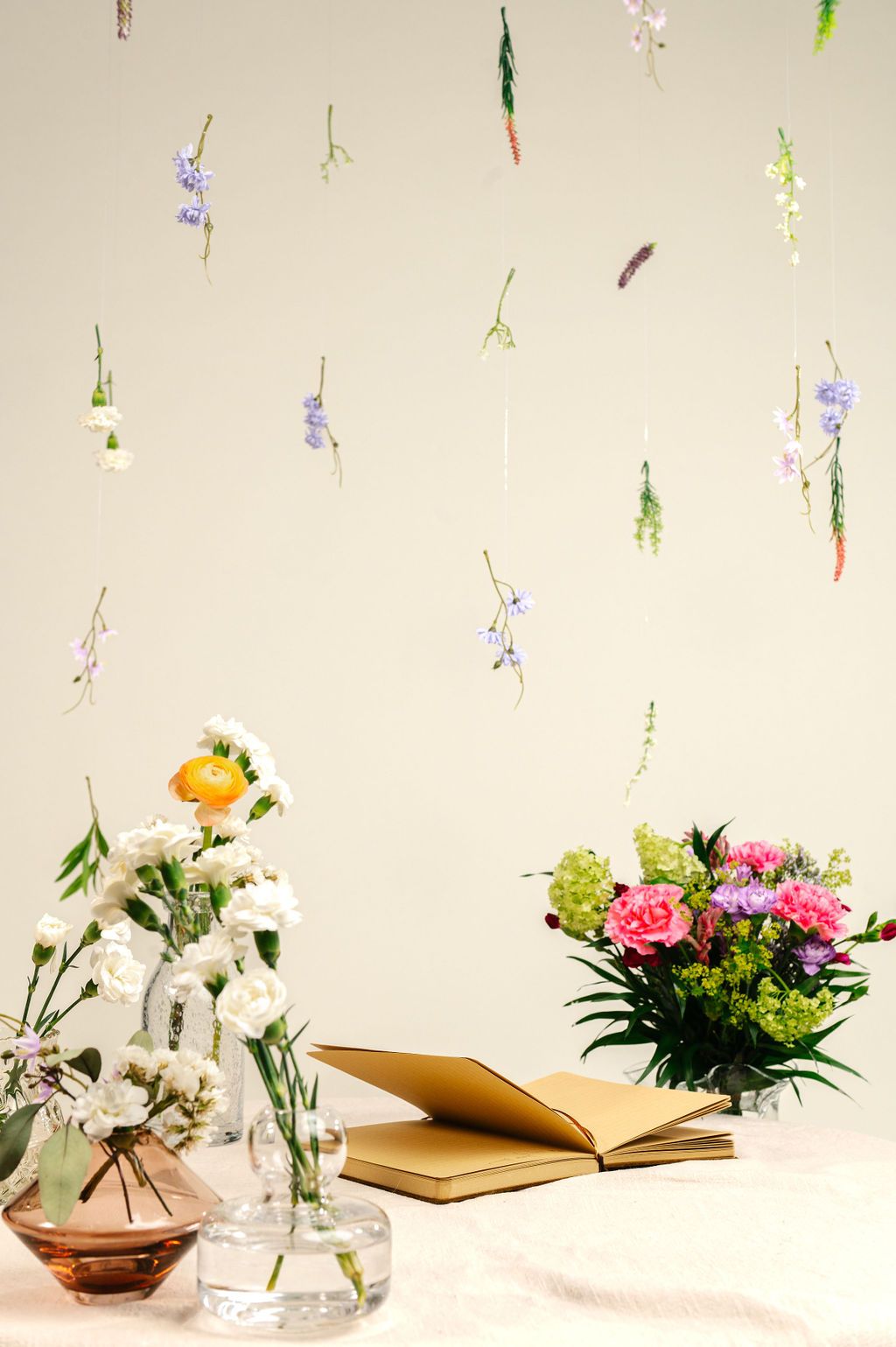 Floral arrangement with vases, open journal, and hanging flowers on a light background.