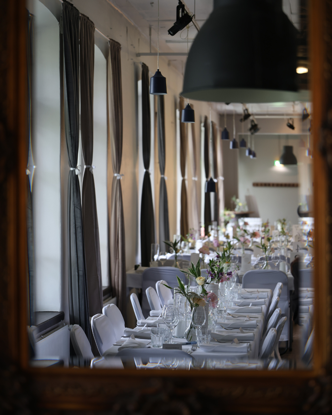 Elegant restaurant dining room with long white-clothed tables, floral centerpieces, gray curtains, and black pendant lights.