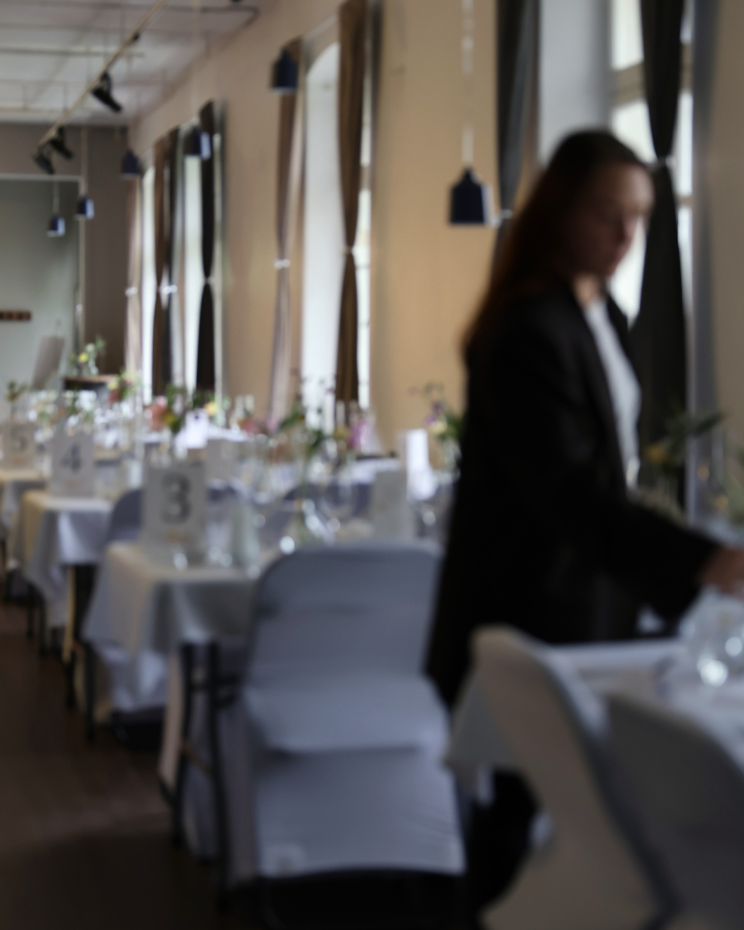 A blurred image of a waitress in black attire walking through an elegantly set dining room with white tablecloths.