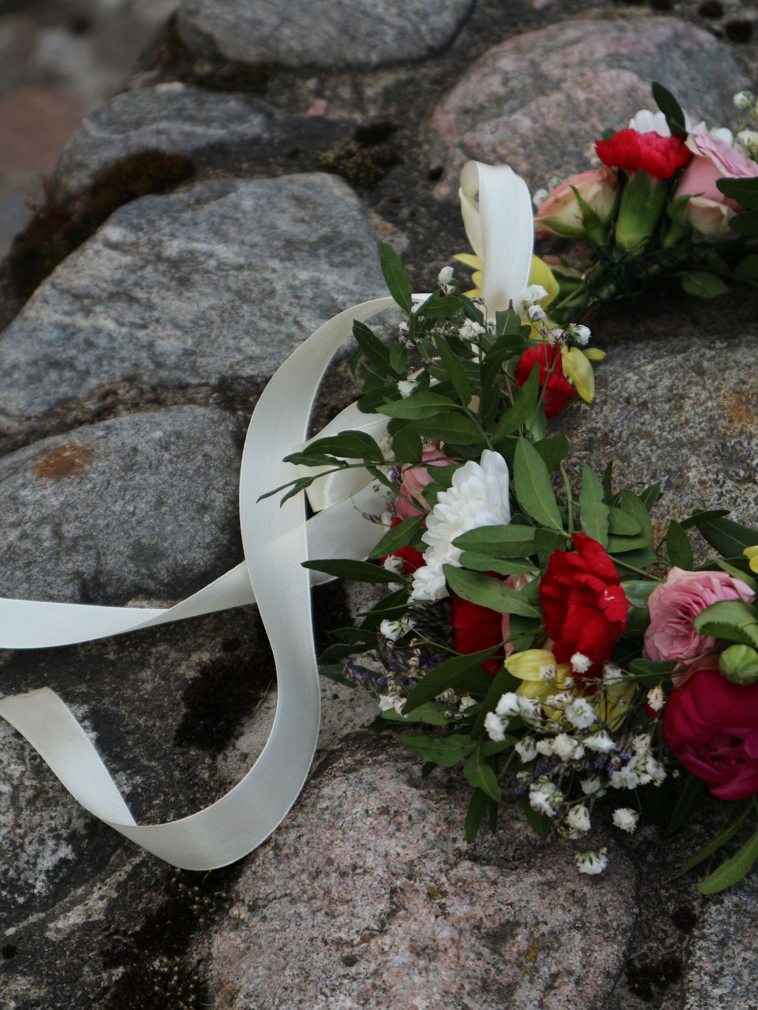 Floral crown with red roses, pink flowers, and baby's breath tied with white ribbon resting on gray rocks.