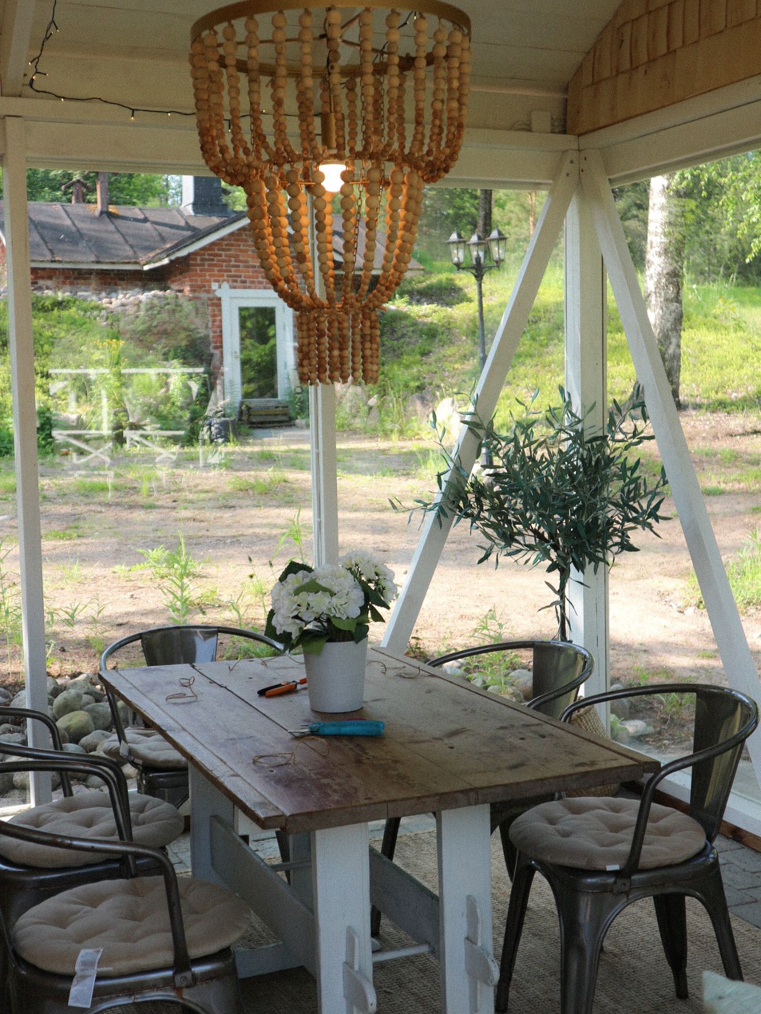 Rustic dining area with wooden table, metal chairs, and beaded chandelier on a screened porch overlooking garden.